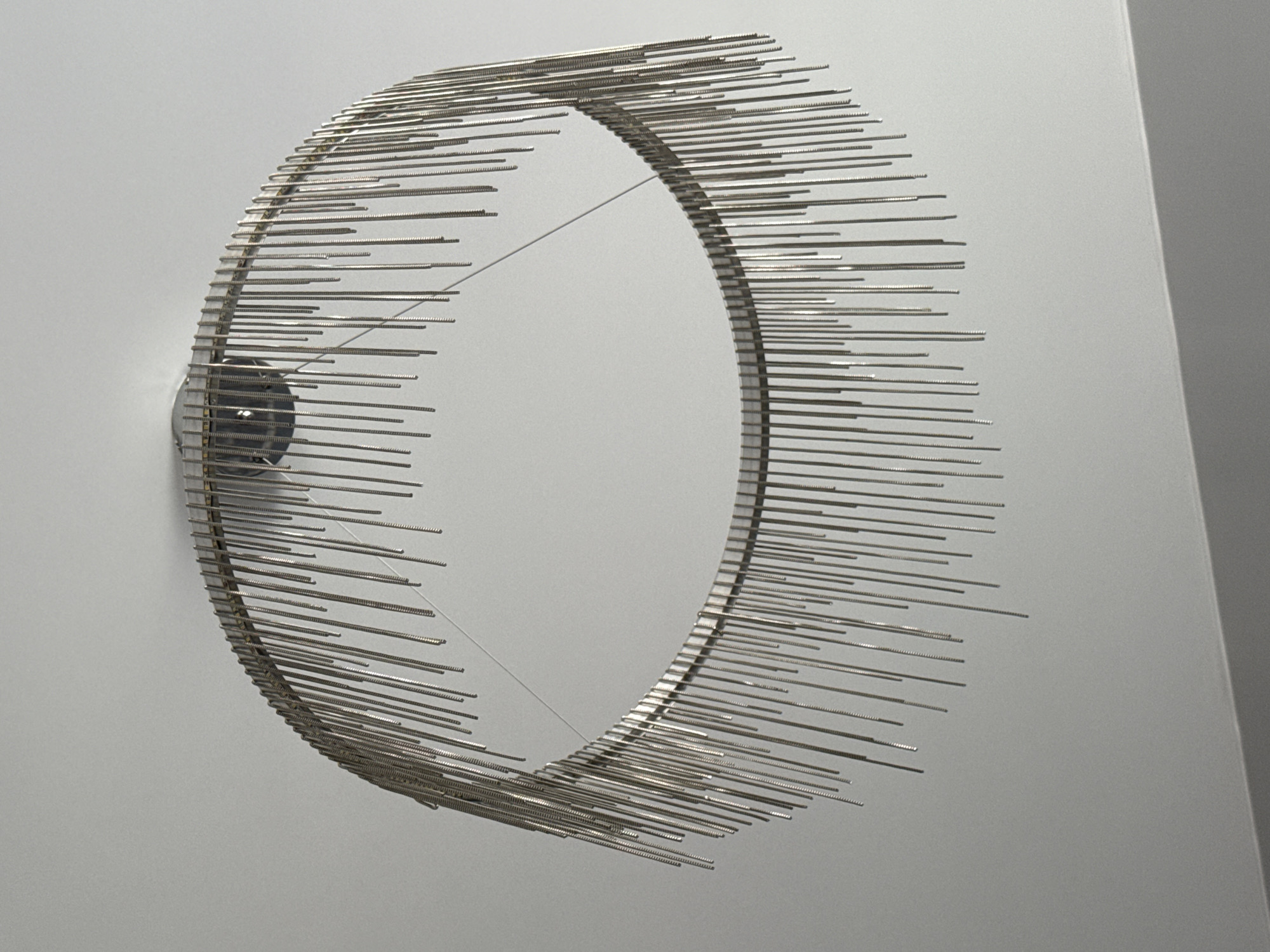 A metal sculpture in the shape of a ring with protruding rods is mounted on a light gray wall. A thin wire crosses the interior of the ring. The location is not discernible from the image.