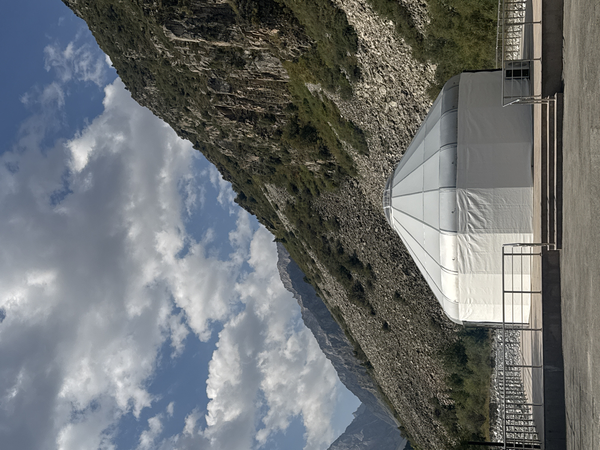 In a mountainous region of Kyrgyzstan, a modern yurt-like structure stands on a raised wooden platform. The yurt features a white fabric base with a translucent, segmented roof. A metal railing encloses the platform, and steps lead up to it. The background is dominated by a steep, rocky mountain slope covered in sparse green vegetation. The sky above is a mix of blue and scattered white clouds. The image appears to be taken on a clear day.