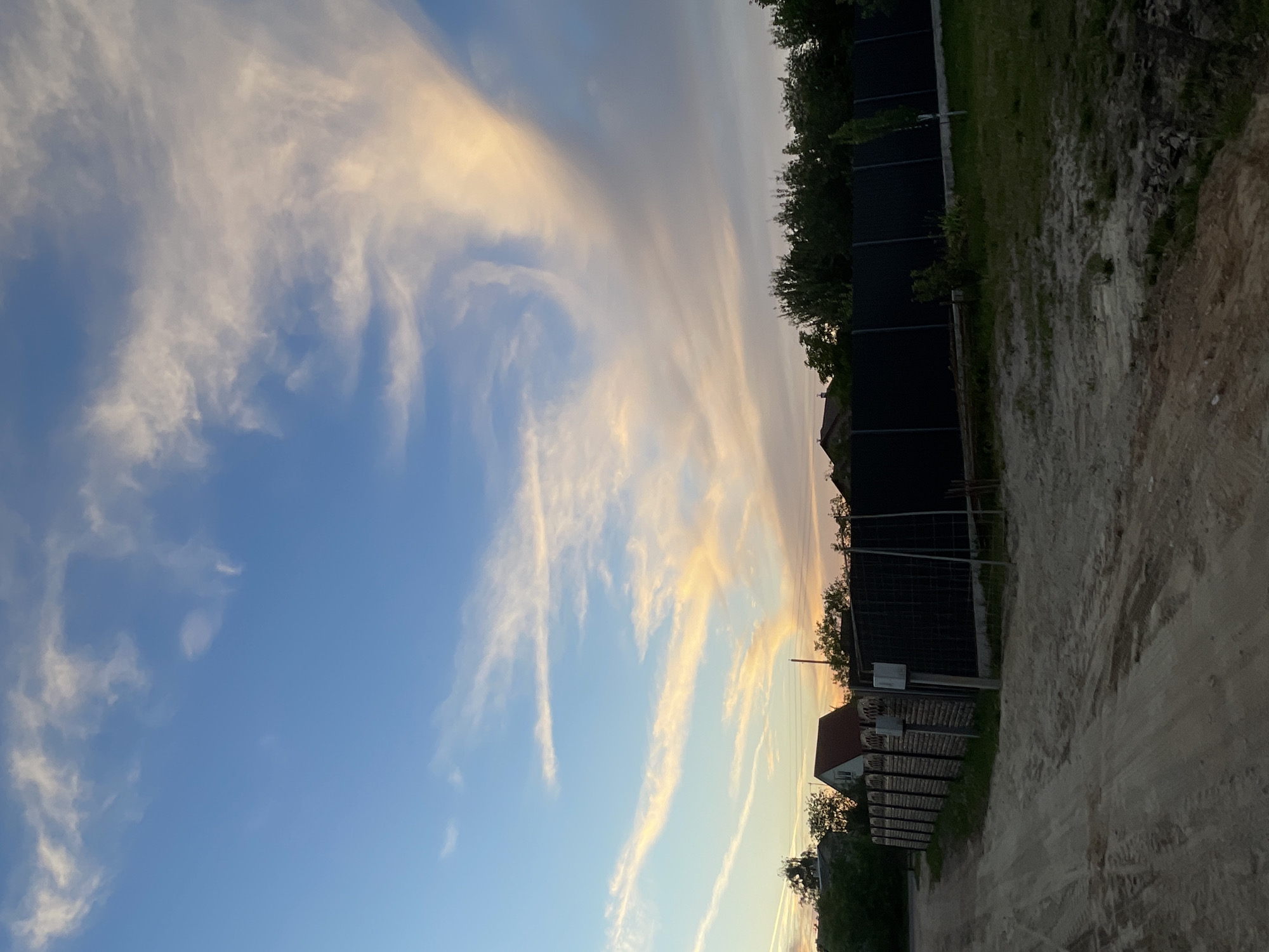 The image depicts a dirt road leading towards a residential area in Мухавецкі сельскі Савет, Belarus, under a sky illuminated by the soft hues of an approaching sunset. The sky is a gradient of clear blue near the horizon, transitioning to a lighter, more pastel shade higher up, with wispy, elongated clouds streaked with orange and yellow, indicating the sun is low in the sky. In the middle ground, a dark, solid fence runs horizontally across the frame, separating the road from the properties beyond. To the left of the fence, a section of a wooden fence with vertical planks is visible, featuring mailbox-like structures attached to posts. Beyond the fences, several houses with red roofs and trees can be seen. The foreground is dominated by the dusty, unpaved road, showing tire tracks and patches of disturbed soil. The overall impression is one of a rural or suburban setting at dusk.