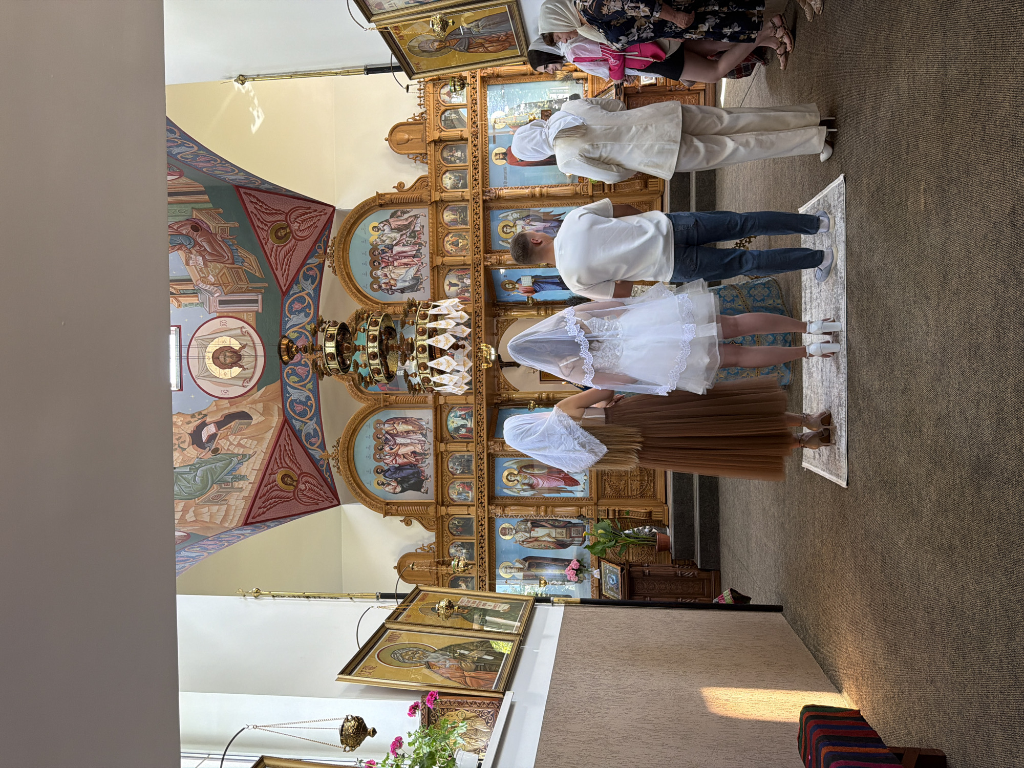 The image depicts an Orthodox Christian wedding ceremony taking place inside a church. In the foreground, a bridal couple stands facing the altar, with the bride in a white dress and veil, and the groom in a white t-shirt and blue jeans. To their left, a woman in a brown dress and veil stands. To the right of the couple, another woman in a cream-colored suit and headscarf stands, and further to the right, two other women in casual attire observe the ceremony. The church interior is adorned with religious icons and murals on the walls and ceiling. A large, ornate chandelier hangs from the ceiling above the altar. The overall scene is one of solemnity and reverence. The location is confirmed to be the Nativity of the Theotokos Church in Strășeni, Moldova.