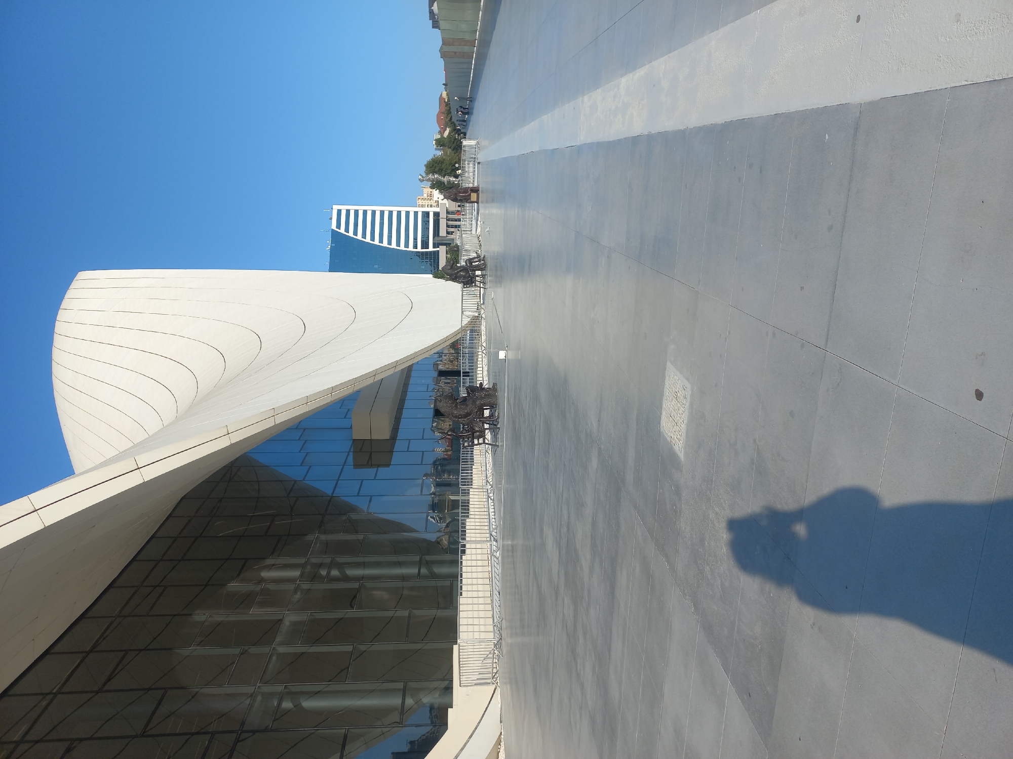 The image shows the exterior of the Heydar Aliyev Center in Baku, Azerbaijan, on a clear day. The iconic, flowing white structure of the center dominates the left side of the frame, with its large glass facade reflecting the sky. In the background, a modern skyscraper with a blue glass exterior and white horizontal stripes stands out against the bright blue sky.
In the foreground, a wide, gray paved plaza stretches out. Several bronze sculptures of people are placed on the plaza, some seated and some standing. A metal railing separates the plaza from the building's entrance. A prominent shadow of a person holding a camera is cast on the plaza in the lower left corner, indicating the photographer's presence. The overall environment is clean and open, with a sense of modern architecture and public space.