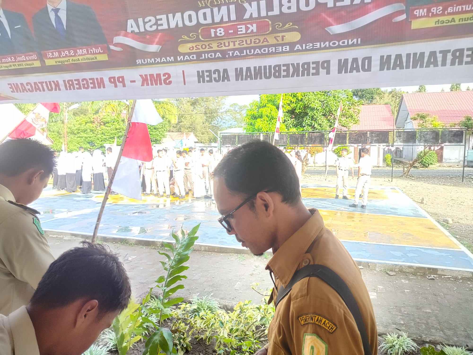 The image captures an outdoor scene in Purwodadi, Indonesia, where a group of individuals, mostly young men in light brown uniforms, are gathered on a paved court marked with blue and yellow lines. A large banner hangs in the background, displaying text in Indonesian and featuring imagery of the Indonesian flag. Some individuals are standing in what appears to be a line, while others are engaged in conversation or observing. The environment includes trees and buildings with red tiled roofs visible in the distance. The overall atmosphere seems to be one of an organized event or gathering.