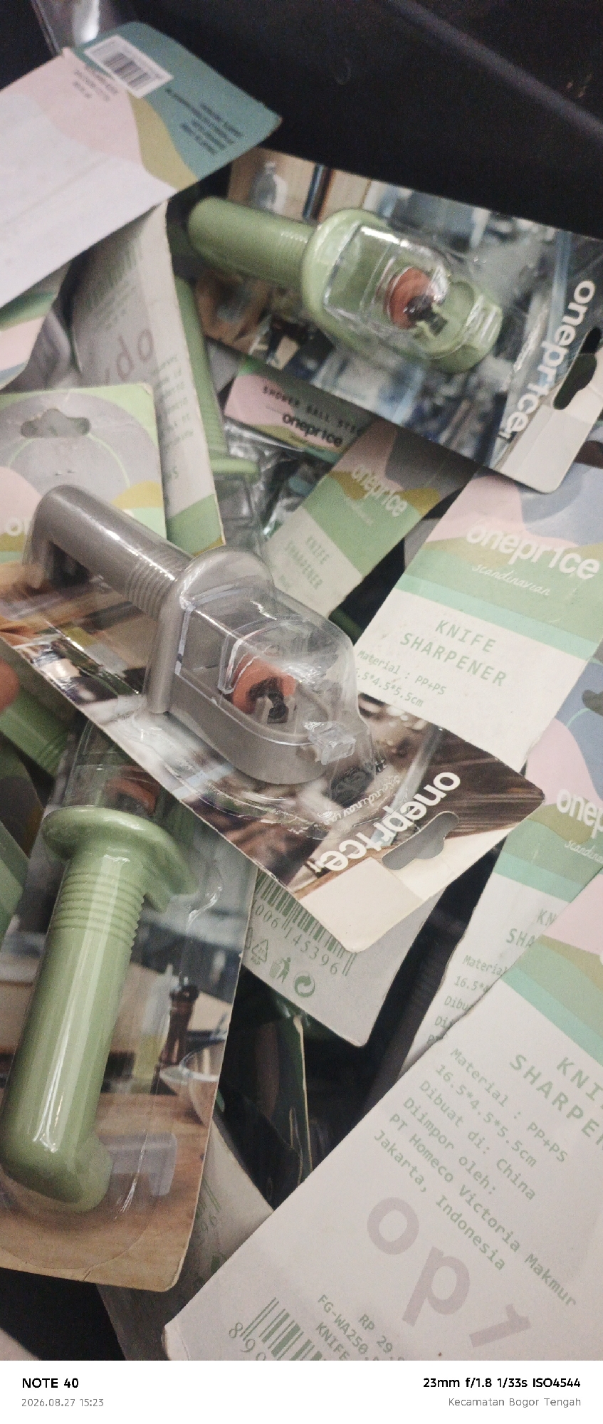 The image shows a close-up view of several packaged knife sharpeners. The sharpeners appear to be of the same model, with a gray handle and a clear plastic casing that reveals a red circular component. The packaging features the "oneprice" logo, and some also include the text "KNIFE SHARPENER" and "scandinavian". Some of the packaging details indicate manufacturing and import information, such as "Material: PP+PS", dimensions like "16.5*4.5*5.5cm", and that it was "Dibuat di: China" and "Diimpor oleh: PT Homeco Victoria Makmur Jakarta, Indonesia". The scene is likely a retail or storage environment, with multiple products stacked and overlapping. The bottom left corner of the image displays text that reads "NOTE 40" and a date and time stamp "2026.08.27 15:23", and the bottom right corner has camera settings "23mm f/1.8 1/33s ISO4544" and the location "Kecamatan Bogor Tengah".