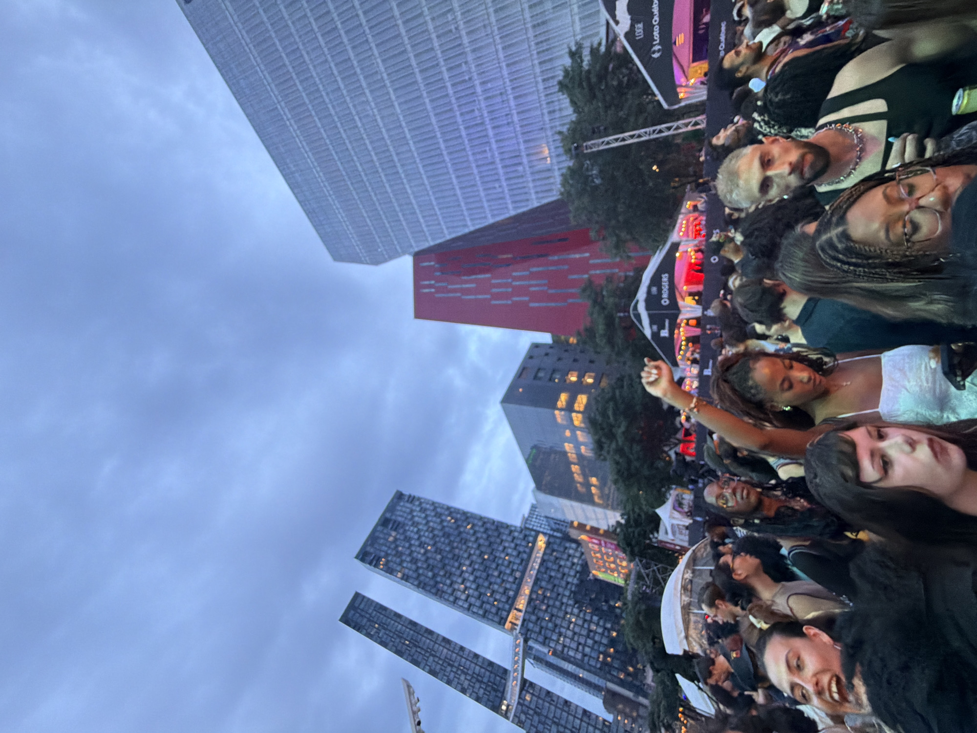 The image depicts an outdoor evening event, likely a festival or concert, in Montreal, Canada. In the background, several modern high-rise buildings are visible. Two prominent dark glass skyscrapers with illuminated windows stand on the left, while a striking red building with vertical window patterns is situated in the middle. A large, light-colored building with horizontal ribbed texture dominates the right side of the frame. The foreground is filled with a crowd of people, suggesting a lively atmosphere. Some individuals are looking at the camera, while others are engaged with the event, with one person in the middle raising their fist. Tents with branding, including "ROGERS" and "Loto Québec," are set up in the mid-ground, indicating corporate sponsorship. The sky is overcast with a blue-grey hue, typical of twilight.