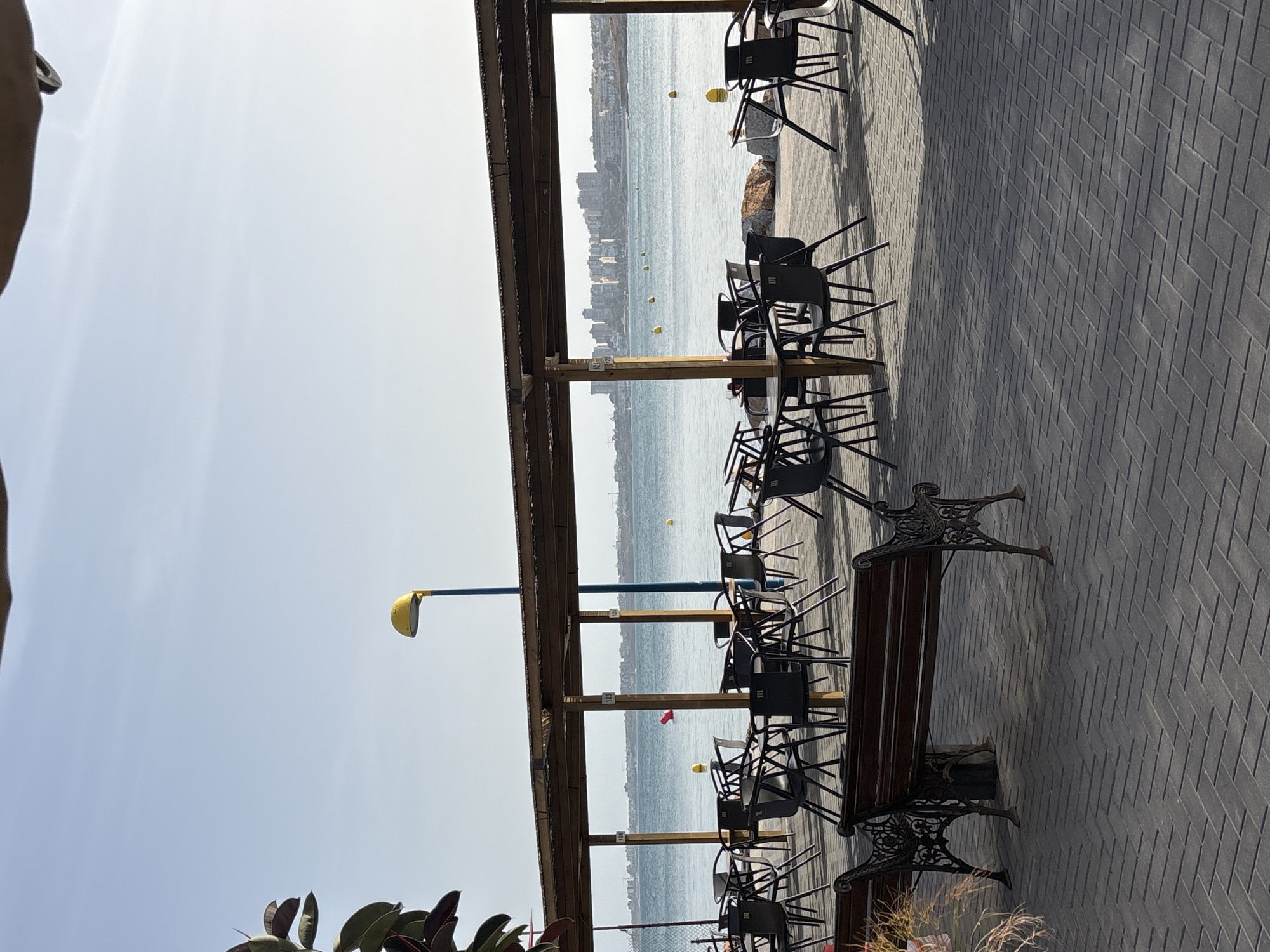 An outdoor cafe with dark plastic chairs and tables is set up on a paved walkway next to a body of water. A wooden overhang covers the seating area, supported by wooden posts. In the background, the water stretches to a distant city skyline under a clear blue sky. A yellow lamp post stands near the water's edge. A dark metal bench with wooden slats sits in the foreground on the paved area.