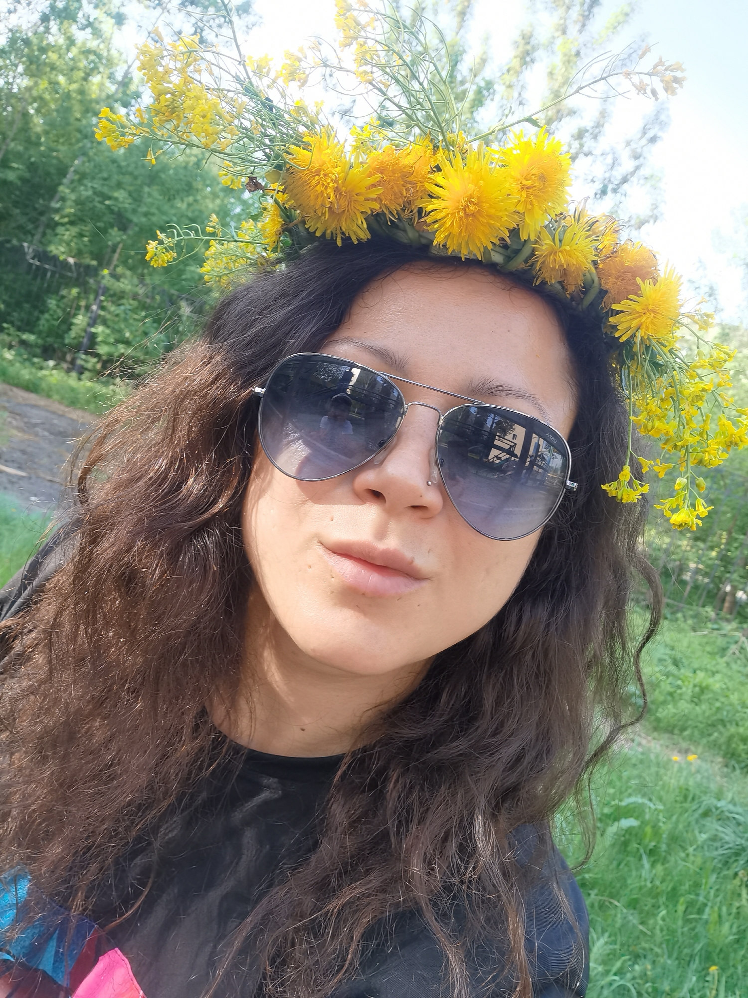 A woman is centered in a medium shot, wearing a crown of yellow dandelions. She is wearing dark-tinted aviator sunglasses and has a small stud piercing in her left nostril. Her dark curly hair falls around her shoulders. She is wearing a dark t-shirt, with a hint of pink and blue design visible on the lower left. The background shows a blurred natural setting, including a patch of green grass with dandelions and trees. The location is in Perm, Russia, and the reflection in the sunglasses appears to show a building from the surrounding environment, with reflective panels and light colored construction.