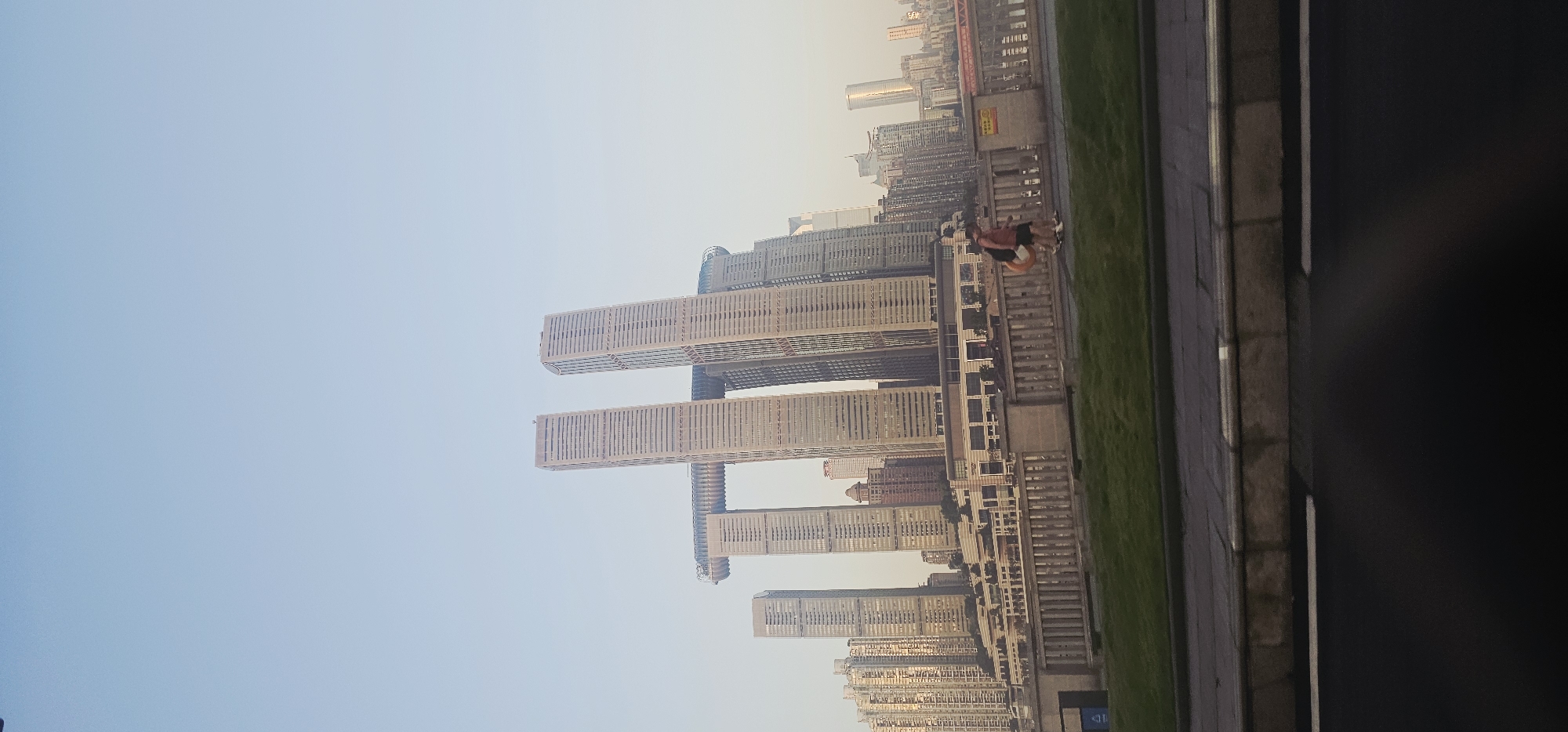 The image shows a view of the Raffles City Chongqing complex in Nan'an District, China, under a pale blue sky. The iconic architecture of the complex, characterized by its twin towers connected by a horizontal structure resembling a ship's hull, is prominent. To the left of Raffles City, a dense cluster of other tall buildings forms the city's skyline. In the foreground, a pedestrian bridge spans across a green grassy area. Two individuals are walking on the bridge, one carrying a large, round, orange object. A street with a dark surface is visible at the bottom of the frame, suggesting the image was taken from a vehicle. The overall scene depicts a modern urban environment.