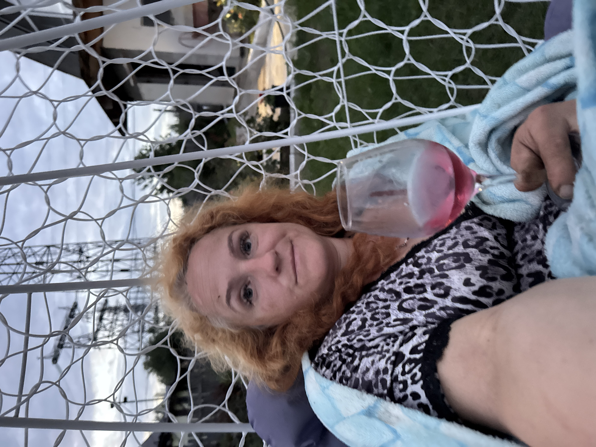 A medium shot shows a woman with red hair sitting in a white net swing chair, wrapped in a blue and white checkered blanket. She is holding a wine glass filled with a pink beverage. She is wearing a black and white animal print top. In the background, a house, trees, and a power line tower are visible under a cloudy sky. The scene appears to be set in a residential area. The location is identified as Chaikovsky, Russia.