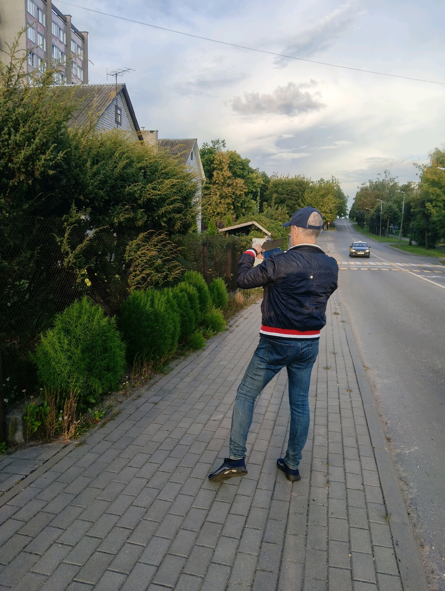 A man in a blue baseball cap, black jacket with red and white trim, and blue jeans stands on a paved sidewalk, holding a phone up as if taking a picture. He is facing away from the camera, towards a road with a car driving on it and trees lining the side. To the left of the man is a chain-link fence, behind which are bushes and a house. Further in the background, a tall, multi-story building is visible. The scene appears to be in a residential area of Hrodna, Belarus, with a cloudy sky overhead.