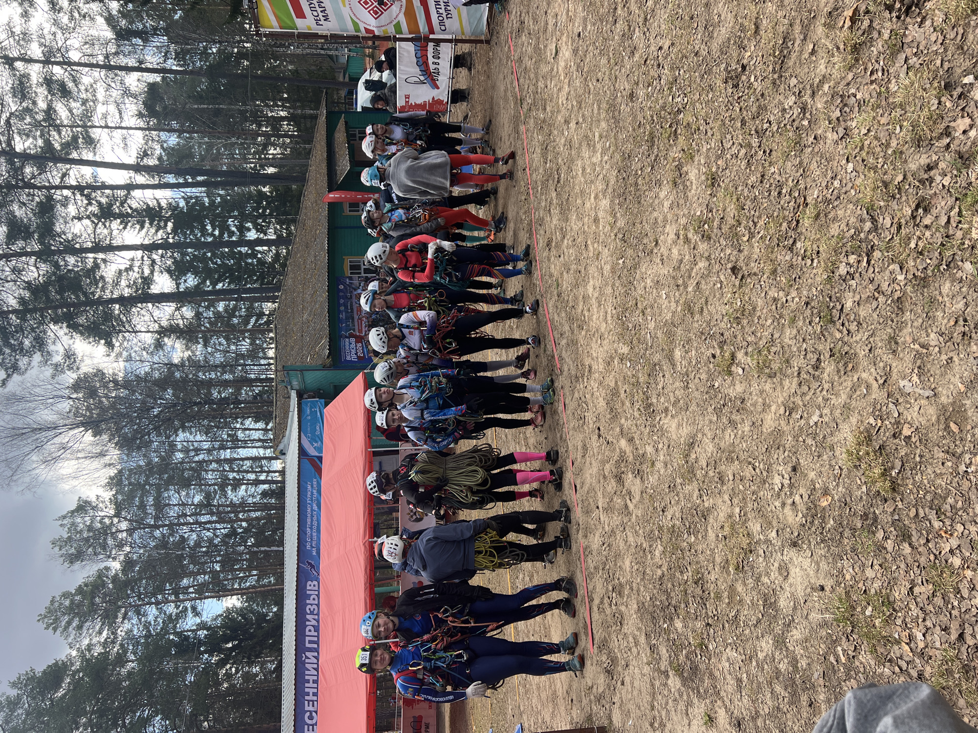 A horizontal photograph shows a line of people wearing climbing gear in Lesnoye, Russia. The people are wearing helmets and harnesses. They are standing on a dirt and leaf covered ground. Behind them is a red tent with white lettering in Russian. Behind the tent is a green building with a brown roof. In the background are tall pine trees. To the right of the people is a banner with the Russian flag and the words "Russia" and "Be in shape."