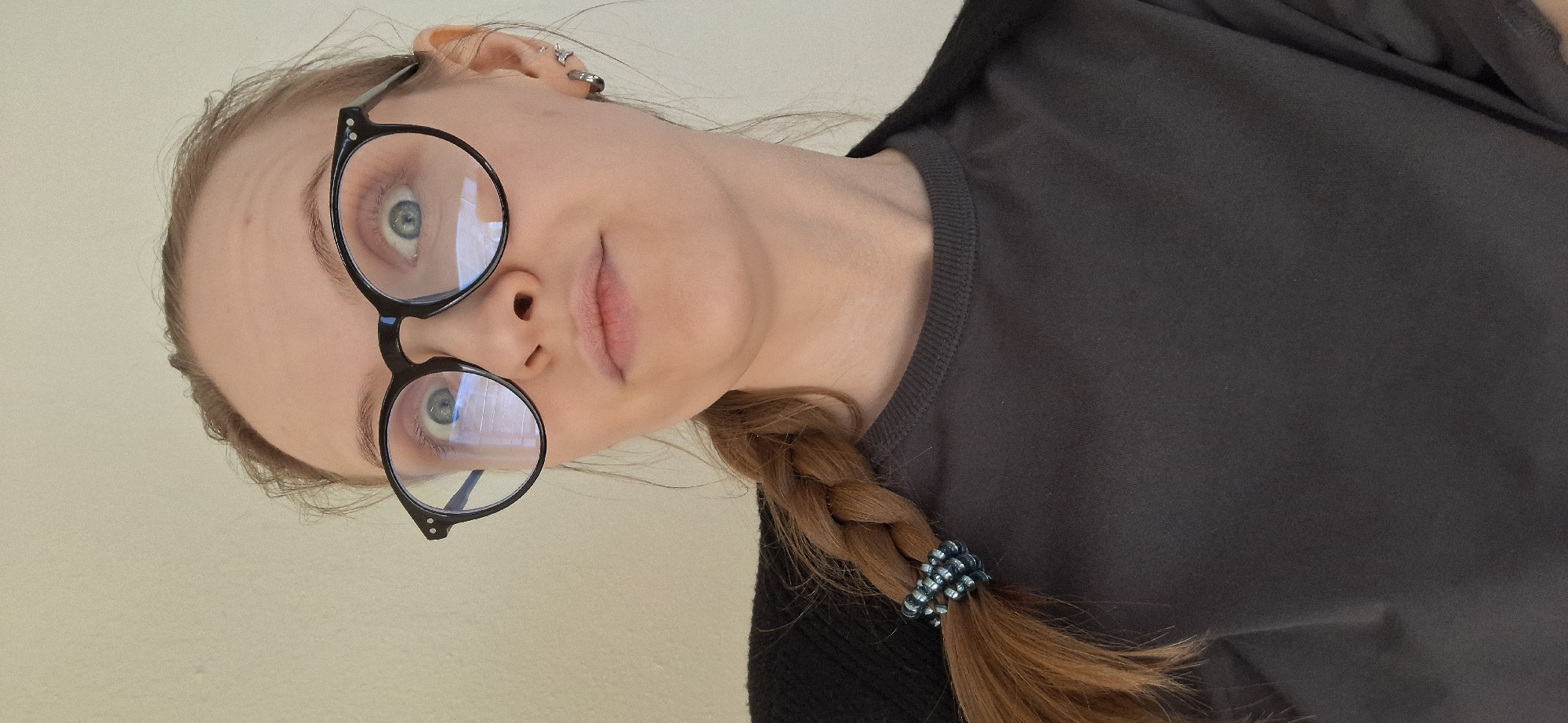 A woman with light skin, wearing round black glasses, is positioned in front of a light-colored wall. She has blue eyes and light brown hair styled in a braid, secured with a beaded hair tie. She is wearing a dark grey t-shirt. The location is not discernible based on the provided image. The city of Chaikovsky, Russia, is not identifiable within the frame.
