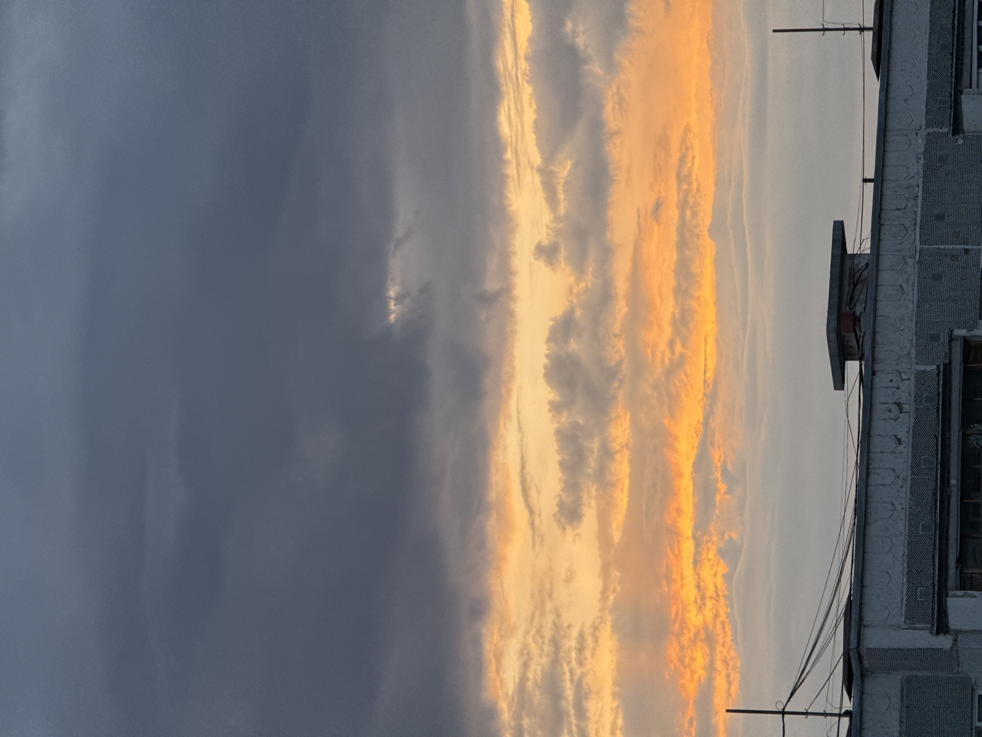 The image shows a portion of a building on the right side, with a cloudy sky occupying the rest of the frame. The building appears to be made of concrete or stone, with rectangular windows and decorative architectural details. Overhead electrical wires extend from the building towards the sky. The sky is divided into two distinct areas: the left side features dark, overcast clouds, while the right side displays orange and yellow hues, suggesting either a sunset or sunrise. The clouds in the center are layered and textured, with a mix of grey, white, and orange tones. The image is rotated 90 degrees clockwise. Without additional context, it's impossible to confirm the specific location within Kazan, Russia.