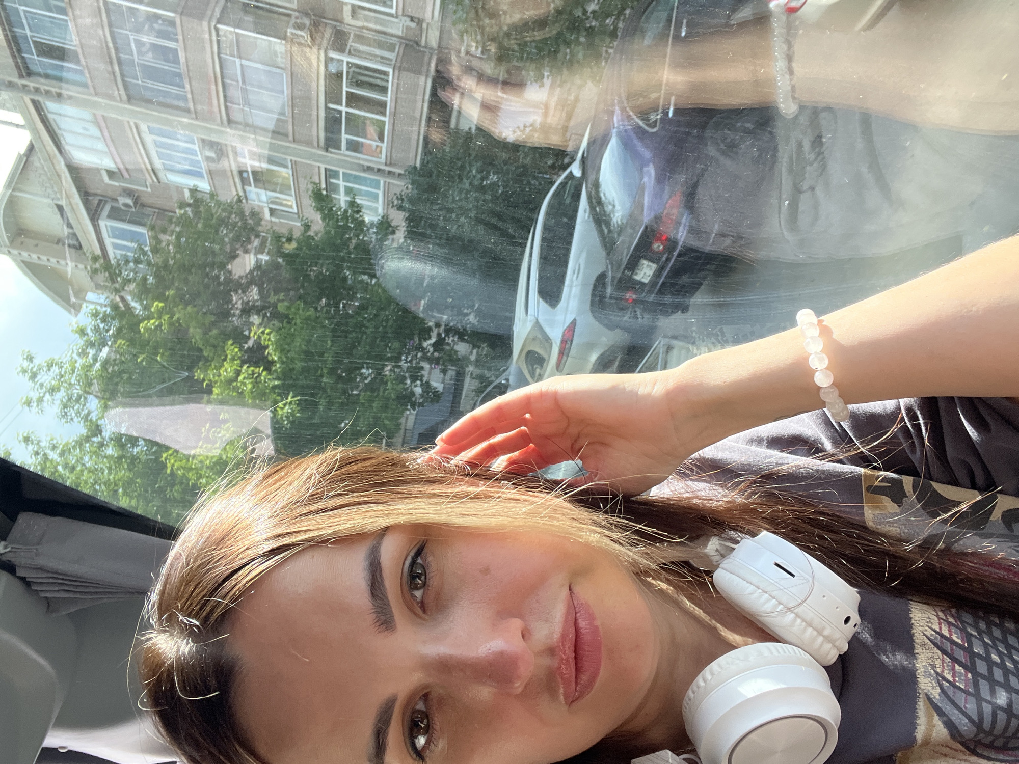 A woman is taking a selfie inside a vehicle. She has brown hair and is wearing headphones around her neck and a bracelet on her wrist. She is looking at the camera and touching her ear. Outside the vehicle, there is a building and cars. The scene appears to be in a city.