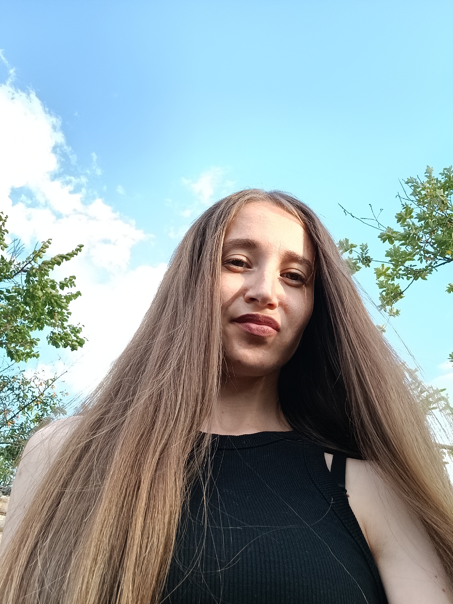 The image is a low-angle, outdoor selfie of a young woman with long, light brown hair. She is wearing a black ribbed tank top. Her head is tilted slightly, and she is looking towards the camera with a neutral expression. The background consists of a bright blue sky with scattered white clouds and green tree branches on the left and right sides of the frame. The lighting suggests it is daytime, possibly late afternoon, with the sun casting shadows on her face. The scene appears to be in a natural, outdoor setting, potentially a park or a garden within the city of Vypasne, Ukraine.