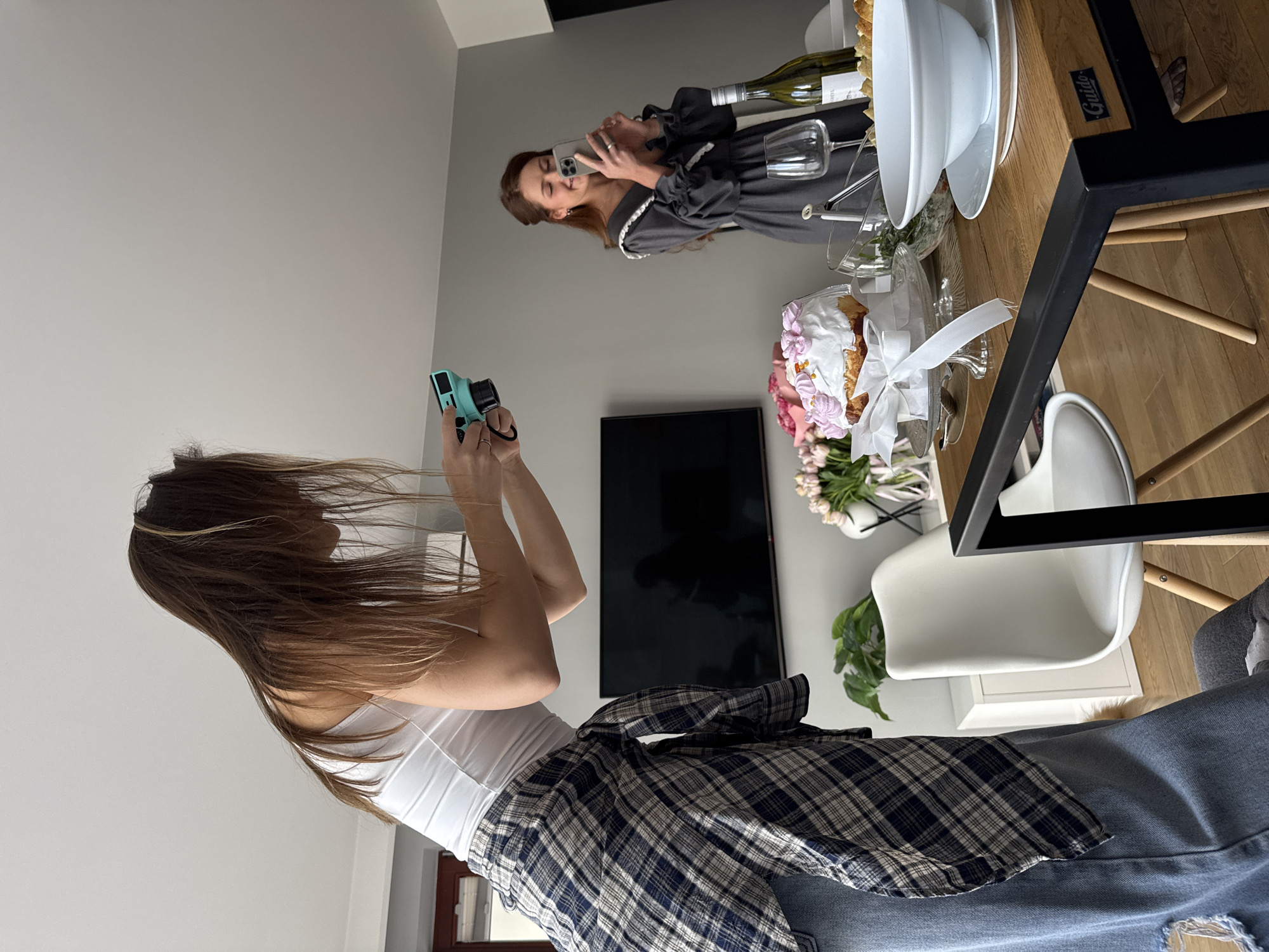 A woman with long brown hair is taking a picture with a light blue camera. She is wearing a white tank top and a plaid shirt tied around her waist, with blue jeans. In the background, another woman is taking a picture with her phone of a table set with food, flowers, and a bottle of wine. A TV is mounted on the wall behind the table. The location appears to be an apartment in Warsaw, Poland.