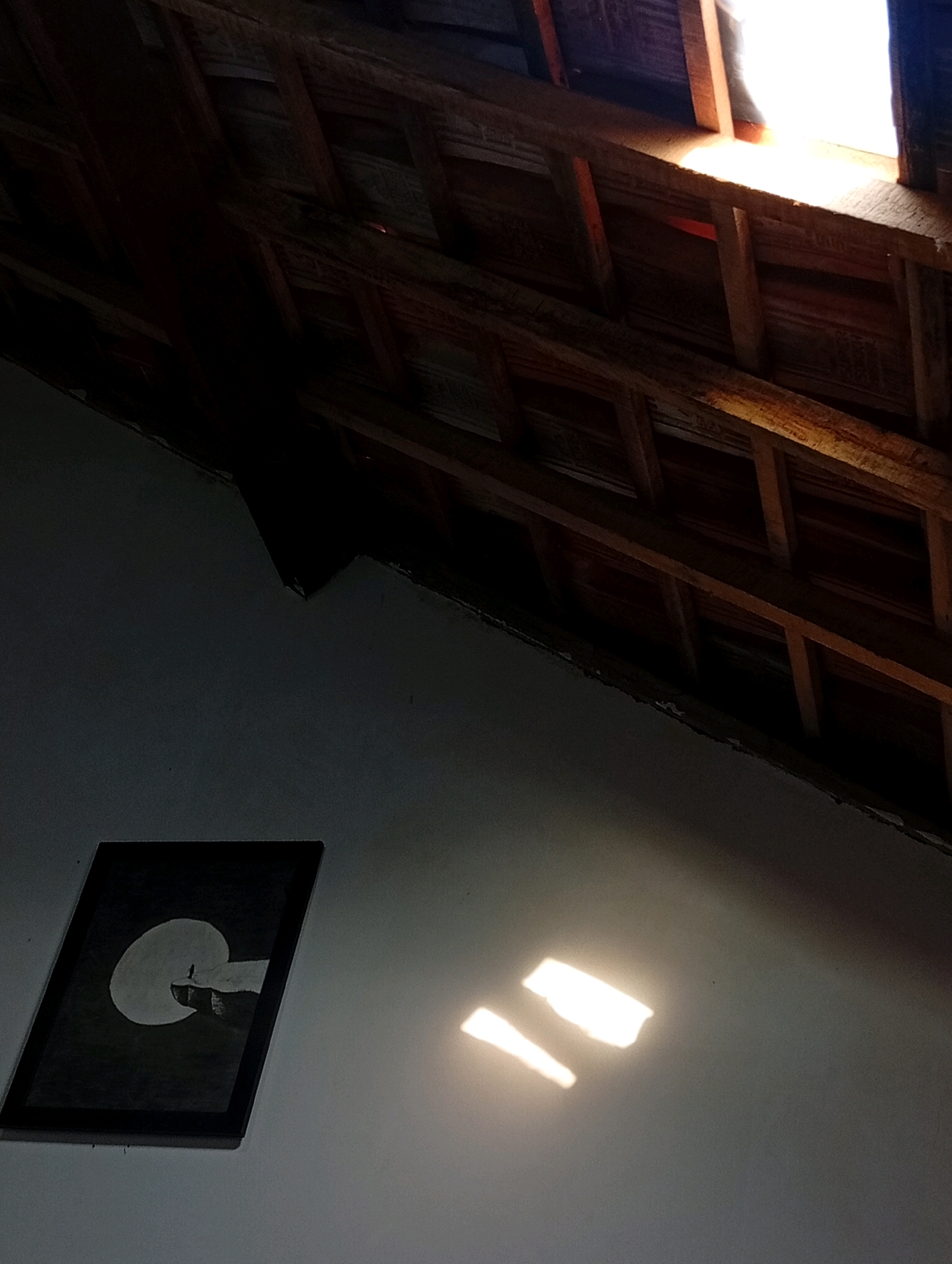 This image captures an interior view of a building, likely a home, in Kebandingan, Indonesia. The scene is characterized by dramatic interplay of light and shadow. The ceiling, constructed with visible wooden beams and planks, is partially illuminated by natural light filtering through an opening, suggesting a skylight or a gap in the roof.
On the wall below, a framed artwork is hung. The artwork depicts a silhouette of a lone figure standing on a ledge, silhouetted against a large circular shape, possibly representing the moon or a full sun. The wall itself is painted in a neutral color and is partially lit by patches of sunlight, creating bright, rectangular reflections. The overall impression is one of rustic simplicity, with the natural light playing a significant role in defining the atmosphere. No people are visible in the image.