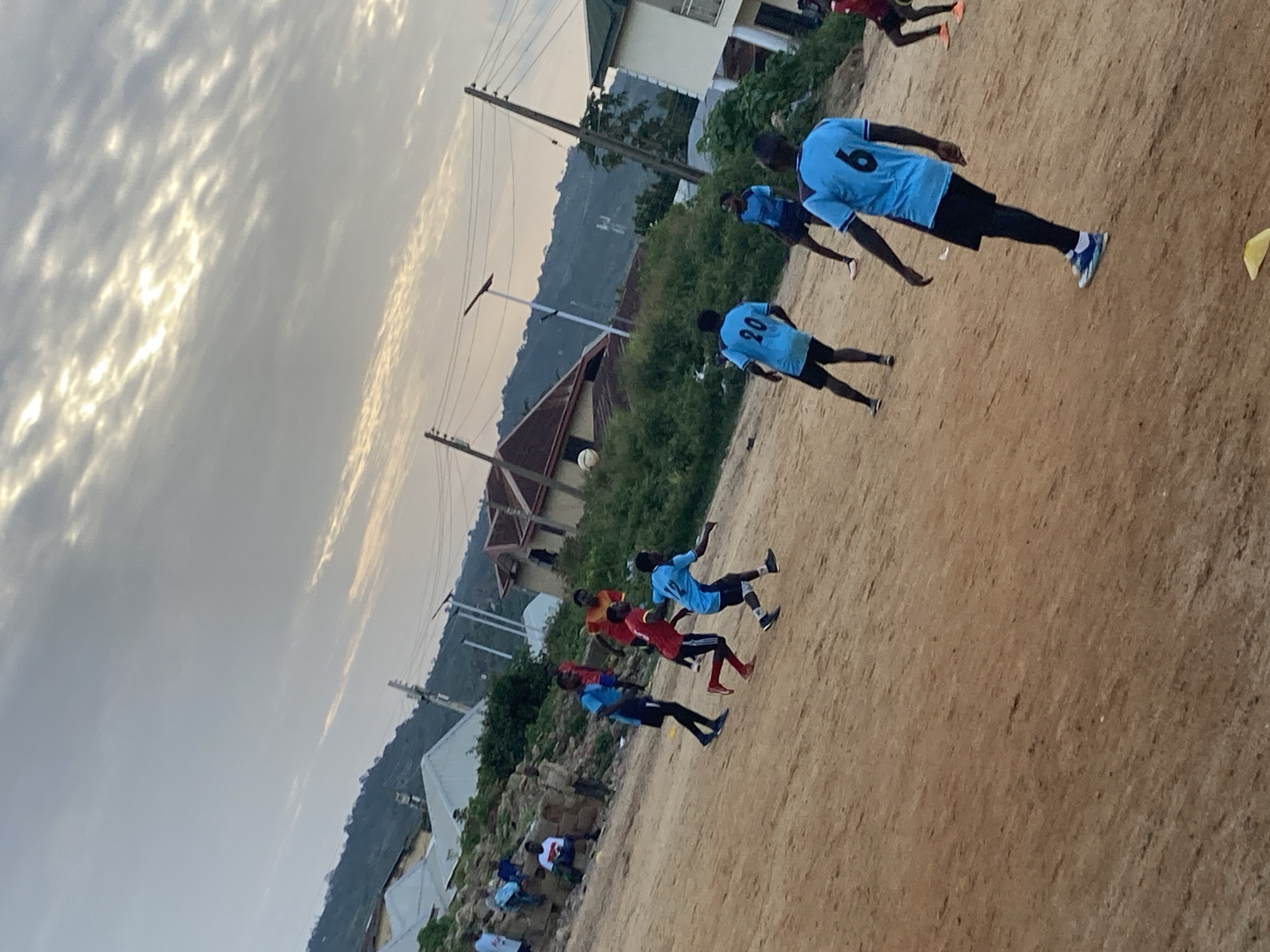 The image depicts an outdoor scene, likely in the city of Jos, Nigeria, where a group of young men are playing soccer on a dirt field. The sky is overcast with some sunlight filtering through the clouds. In the background, there are residential buildings, some with corrugated metal roofs, and hills covered in vegetation.
Several individuals are actively engaged in the soccer game. Some are wearing blue jerseys with numbers on the back, while others are in red jerseys. One player in a blue jersey with the number "6" is prominently featured in the foreground, with his back to the camera. Another player in a blue jersey with the number "20" is also visible. A soccer ball is in mid-air, indicating the game is in progress.
The environment appears somewhat rural or undeveloped, with the dirt field characteristic of many informal playing grounds. Power lines and utility poles are visible in the background, suggesting the presence of infrastructure in the area. Some spectators are seated or standing in the background, observing the game. The overall impression is a casual, community-based sporting activity taking place in a residential area.