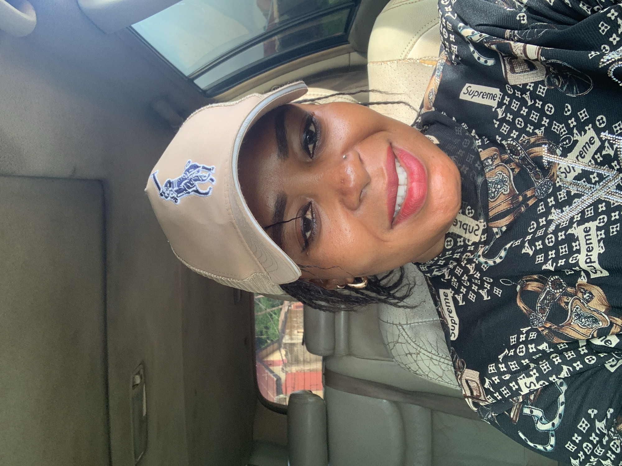 A portrait-style image, taken inside a car in Sabon Tasha, Nigeria, shows a person smiling directly at the camera. The subject, likely a woman with dark skin, is positioned slightly off-center, with her head tilted, suggesting a selfie or a casual snapshot. She wears a light-beige cap with a blue embroidered logo resembling the Ralph Lauren Polo rider. Her dark hair is visible beneath the cap, and she has a small nose piercing and a gold hoop earring. Her face expresses happiness, with a wide smile revealing white teeth and lips colored with red lipstick.
She is dressed in a dark-colored top featuring a repeating pattern that includes "Supreme" text, Louis Vuitton-style monogram elements (LV initials and floral motifs), and equestrian-themed imagery such as belts, stirrups, and horseshoes. The car's interior is visible, with light-colored, possibly leather, seats and a matching headliner. A sunroof is present above. Through the car window, an outdoor scene is visible, showing distant buildings and greenery under what appears to be bright daytime light, suggesting sunny weather. The overall impression is a cheerful moment captured inside a vehicle during the day.