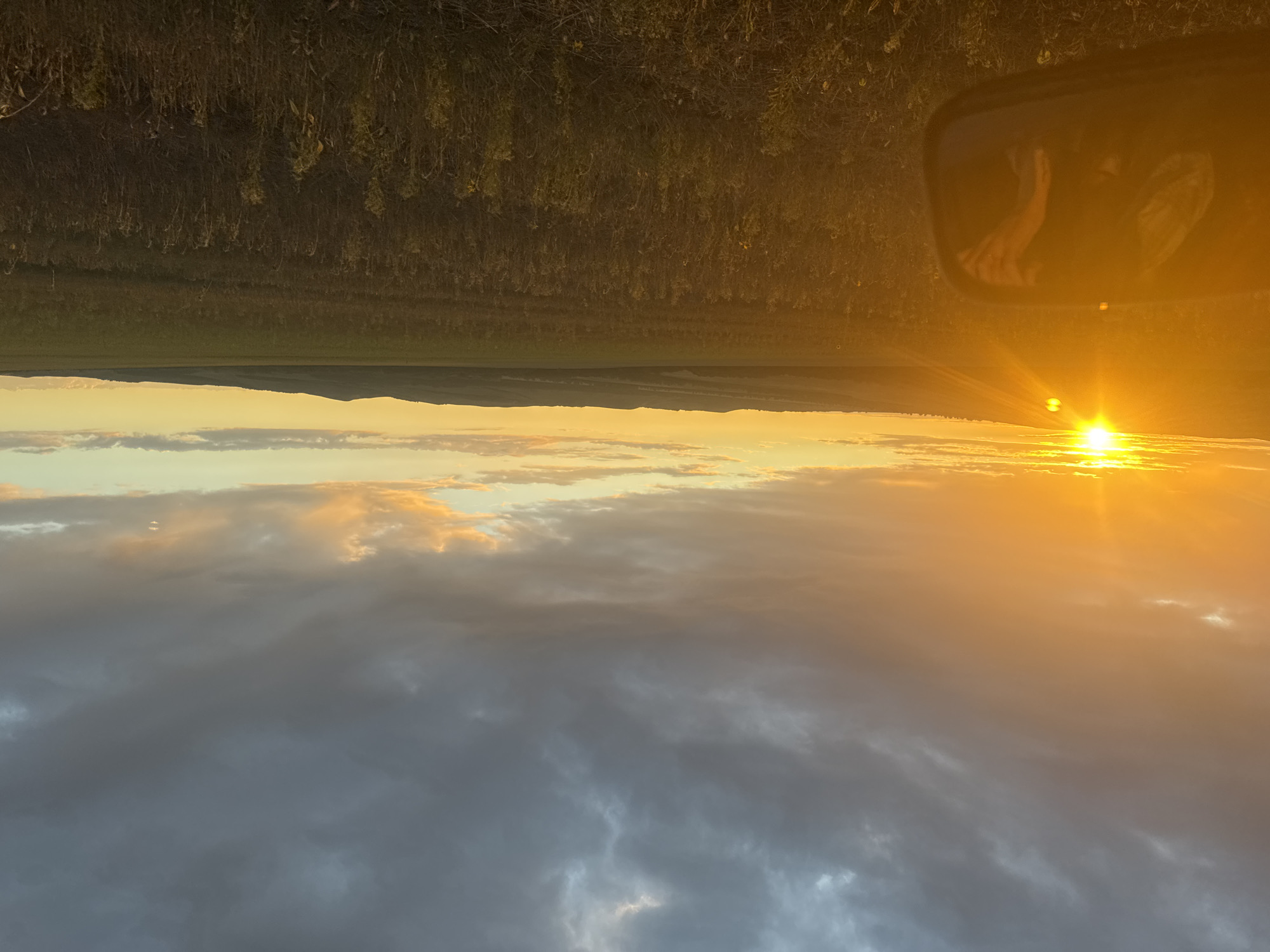 The image captures a wide, rural landscape at sunset, as viewed from inside a vehicle. The sun, a bright orange orb, is low on the horizon to the left, casting a warm glow across the sky and illuminating the scene with a golden light. The sky is a mix of dramatic, dark gray clouds overhead and softer, lighter clouds tinged with orange and yellow closer to the horizon. In the foreground, a side-view mirror of the vehicle is visible on the left, reflecting a blurry image of what appears to be a person's arm and hand. Beyond the mirror, a vast field of dry, tall grass and some scattered yellow flowers stretches towards rolling hills in the distance. The overall impression is one of tranquility and the end of a day in an agricultural area. The location is specified as the city of Khutorka, Russia.