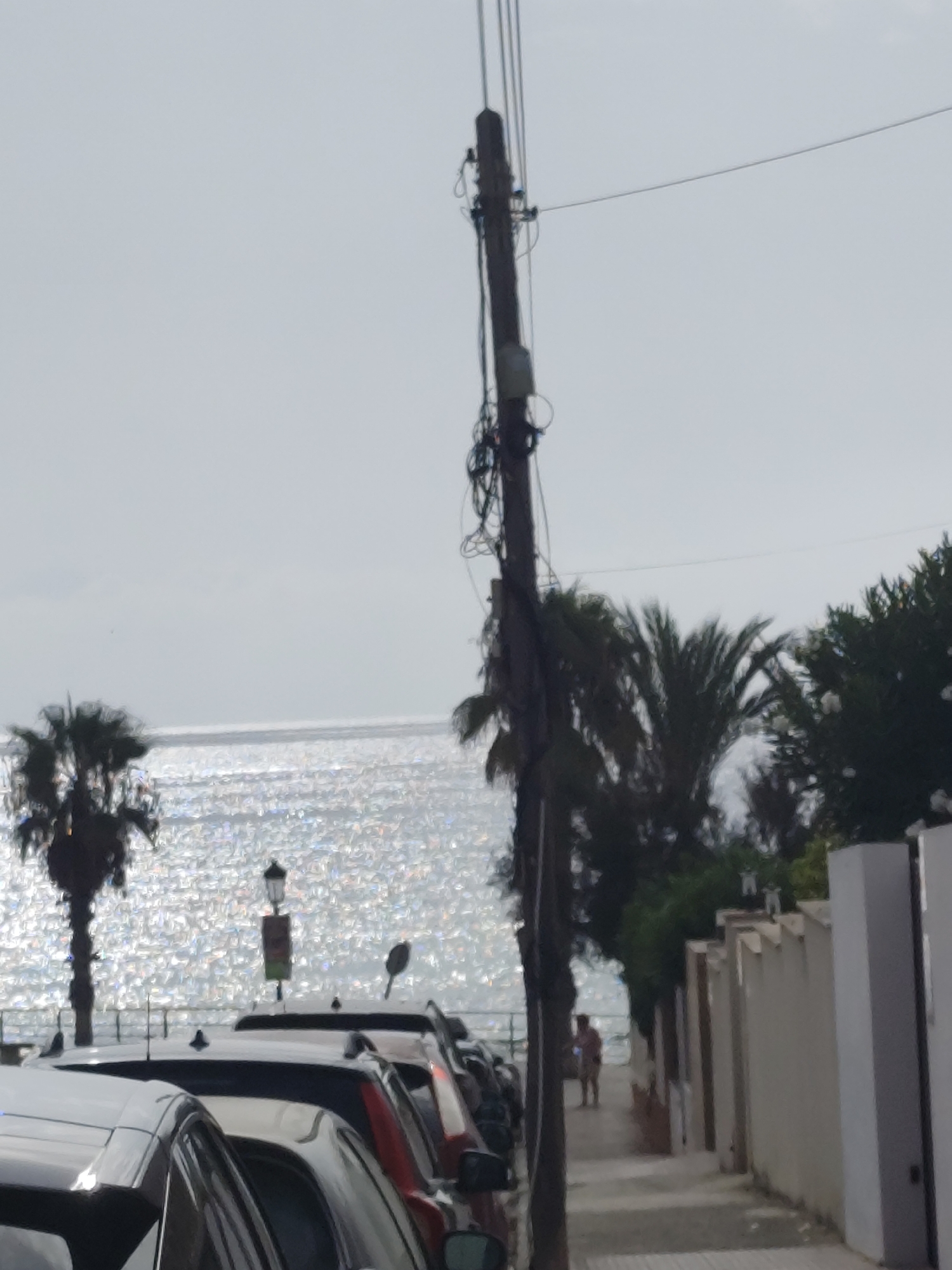 This is a photograph taken outdoors, likely during the daytime due to the bright sunlight reflecting off the water. The scene depicts a waterfront area with several parked cars in the foreground, suggesting a street or parking area adjacent to the coast. Beyond the cars, the ocean is visible, with its surface shimmering intensely, indicating a sunny day with potentially small waves or a breeze.
On the left side of the image, silhouetted palm trees are present, a common feature in coastal or tropical locations. In the middle ground, a lamppost with a sign is situated near the water's edge, and further down the pathway, a person can be seen walking away from the camera. The background features more vegetation and a white wall, possibly part of a building or fence, with several doorways or openings.
A prominent element in the image is a tall utility pole with numerous wires and some equipment attached to it, positioned centrally and extending upwards towards the sky. The sky itself is overcast or hazy, with a light gray tone. The overall impression is a casual, everyday scene at a seaside location. There is no visible text in the image.