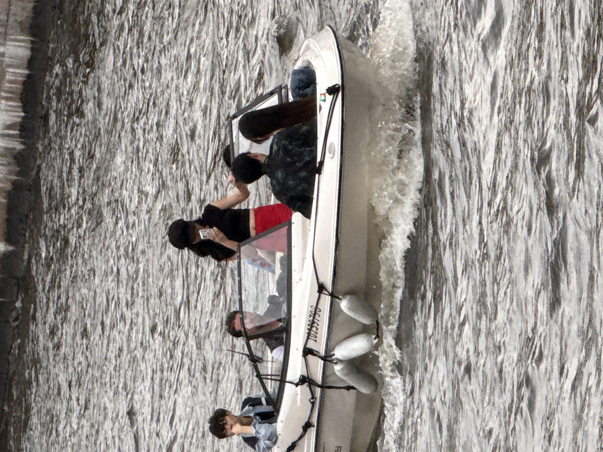 The media file shows a boat with four people on a body of water. The boat is white with a windshield and appears to be moving, creating a wake. The people are positioned in different parts of the boat. One person, wearing a black shirt with a camouflage pattern, is seated in the back, facing forward. Another person, wearing a black beanie and black shirt with red shorts, is standing and looking towards the front of the boat. A third person, wearing a black beanie and dark clothing, is also in the boat. The fourth person, wearing a light-colored shirt, is seated at the front of the boat, looking towards the right.
The water is choppy and appears to be a river or canal, given the context of Saint Petersburg. The lighting suggests it is daytime, with some glare on the water, indicating a bright day. The overall scene depicts a recreational activity, likely a boat ride. There is visible text on the side of the boat, which appears to be a registration number or identifying mark. The image's orientation is rotated, with the boat moving from left to right in the frame.