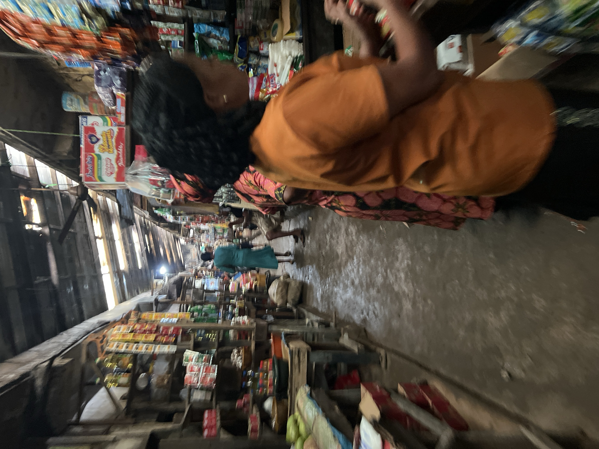 A busy indoor market in Nkpor, Nigeria, is depicted, with a narrow aisle lined with stalls displaying various goods. The aisle floor is wet and appears to be made of concrete. On the left, shelves are laden with packaged food items, bottles of oil, and what appear to be produce. On the right, a woman in an orange t-shirt is partially visible, facing away from the camera and seemingly engaged in an activity at a stall. Behind her, another woman in a floral dress is standing. Further down the aisle, another person in a teal dress is walking away, and two individuals are seated. Overhead, a corrugated metal roof with visible beams and lighting fixtures suggests a covered but open-air structure. The overall impression is one of a vibrant and active marketplace.
