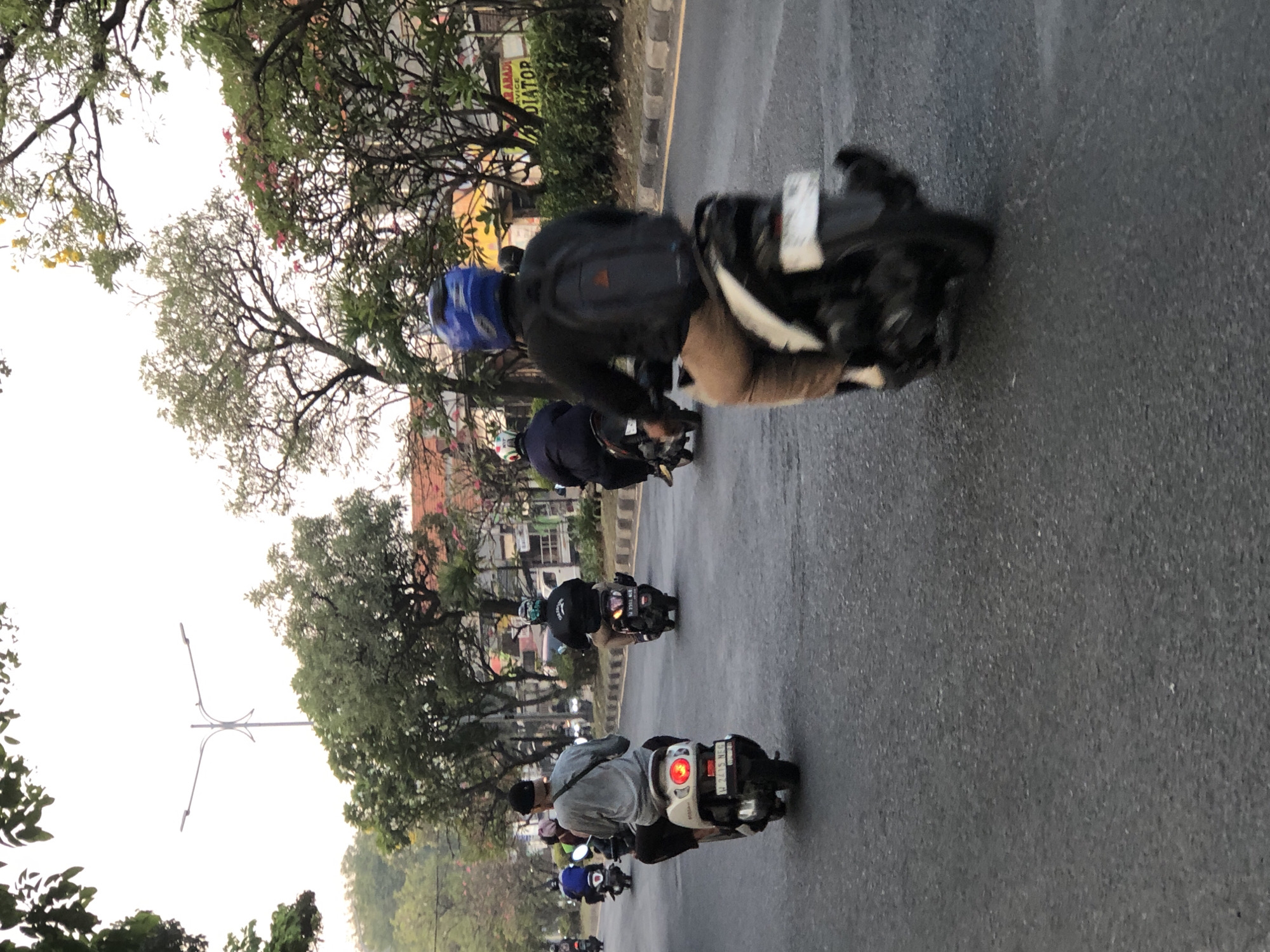 The image captures a street scene in Sumorame, Indonesia, where several individuals are riding motorcycles. The perspective is from behind a person on a motorcycle, looking forward. In the foreground, a person wearing a black jacket, tan pants, and a blue helmet is riding a black motorcycle. Further ahead, another person in a gray shirt and black pants, also on a white motorcycle, is visible. The road is paved, with a yellow line marking the lane. Trees line the side of the road, with some buildings and a street lamp in the background. The sky is overcast. The overall atmosphere appears to be typical of daily life in an Indonesian city.