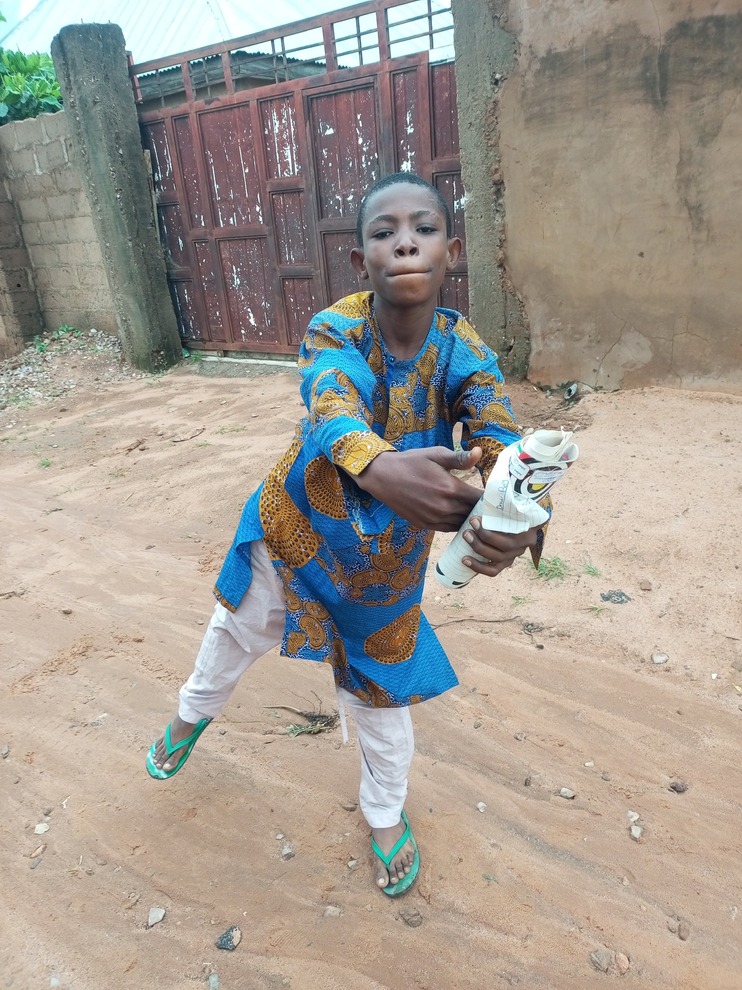 A young boy stands in a dirt area, holding a rolled-up piece of paper or fabric in his hands. He is wearing a blue and gold patterned shirt and white pants, along with green flip-flops. In the background, there is a large, weathered metal gate and a rough, sandy wall. The ground is covered in sand, small rocks, and some sparse vegetation. The scene appears to be outdoors, possibly in a residential area or a less-developed part of a city.