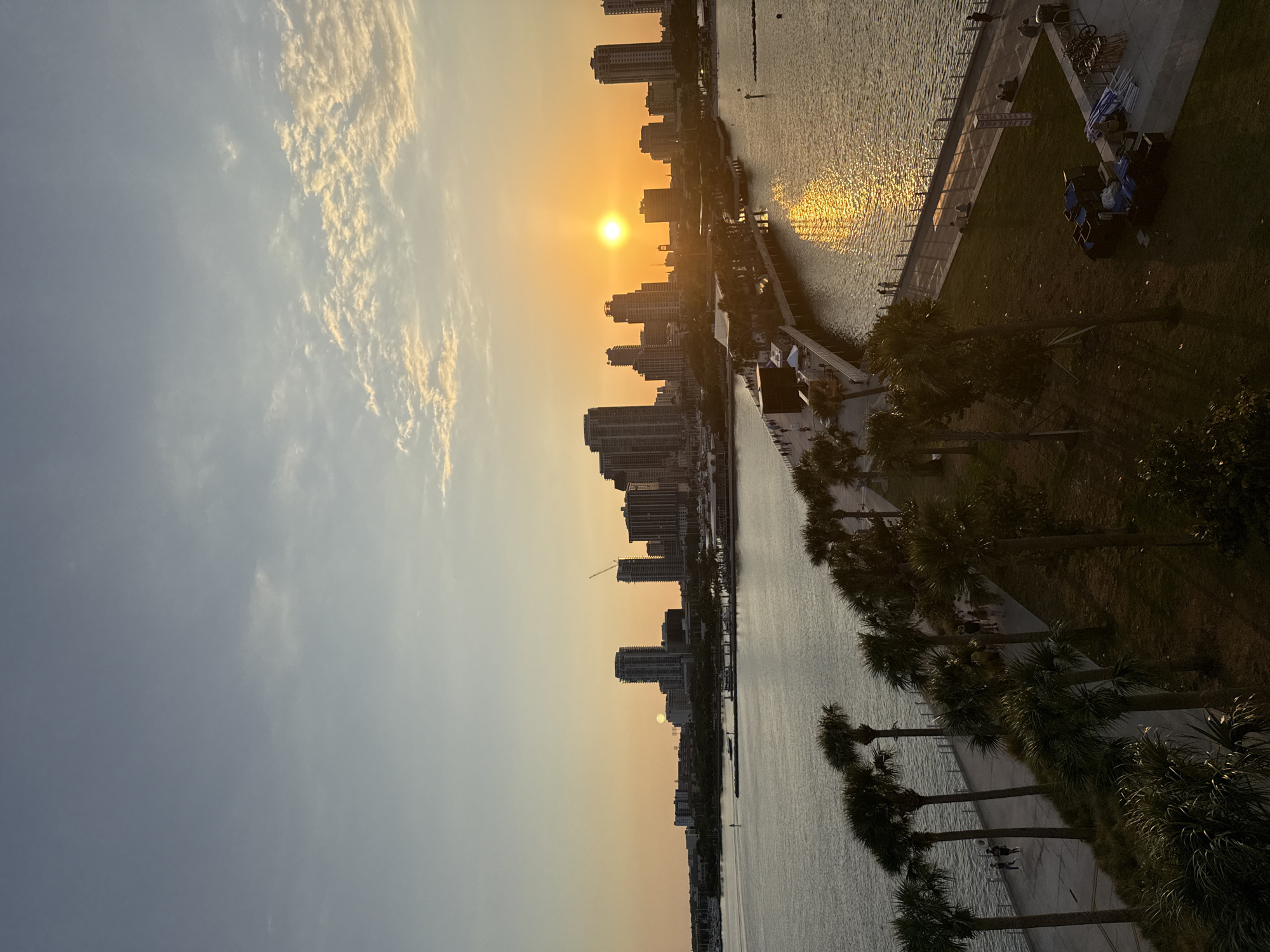 An elevated, wide-angle image depicts the waterfront and downtown skyline of Saint Petersburg, Florida, United States of America, at sunset. The sky transitions from a light blue at the top to a vibrant orange and yellow hue near the horizon, with the sun prominently visible as a bright, circular, orange orb above the city buildings. Wispy clouds are scattered in the upper sky.
The cityscape in the background consists of numerous high-rise buildings and skyscrapers, several appearing as dark silhouettes against the bright horizon. A construction crane is visible atop one of the taller structures. To the far left, a marina populated with multiple boats is visible.
In the middle ground, an expansive body of water, likely Tampa Bay, reflects the setting sun, creating a shimmering golden path extending from the sun towards the foreground. The St. Pete Pier extends from the land into the water on the right, curving slightly to the left. It features various structures, including larger buildings further out and smaller kiosks or tents closer to the land. Numerous individuals are visible walking along the pier. A white navigational buoy is present in the water to the right of the pier.
The foreground features a strip of land with a manicured grassy area and numerous tall palm trees lining a paved waterfront promenade. People are observed walking along the promenade and seated on the grass. On the far right, on a paved surface adjacent to the grass, various items resembling event equipment, including stacked boxes, tarpaulins, tent poles, and bicycle racks, are present. The overall lighting indicates the golden hour, casting long shadows and highlighting silhouettes.