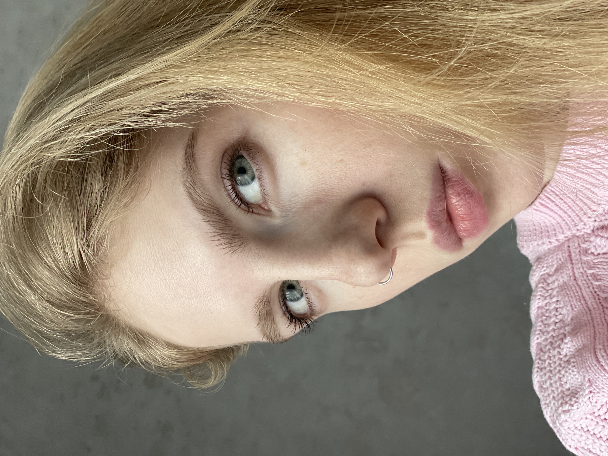 A close-up portrait shows a young individual, likely female, with fair skin and light blonde hair. The image is oriented with the subject's head tilted, focusing on the upper part of the face. The subject's light blue-grey eyes are prominent, looking slightly upwards and to the right within the frame. Long, dark eyelashes frame the eyes.
A small, silver hoop ring pierces the left nostril. The lips are full, a soft pink color, and appear slightly parted and glossy. Blonde hair, appearing fine and light, frames the forehead and sides of the face. Part of a light pink, possibly knitted, garment is visible on the right side of the image, suggesting the subject is wearing a sweater or similar top.
The background is a plain, dark grey color, providing a neutral setting that highlights the subject. The lighting is soft and even, without harsh shadows, emphasizing facial features and skin texture. No specific activity is depicted beyond posing for the portrait. There is no visible text in the image.