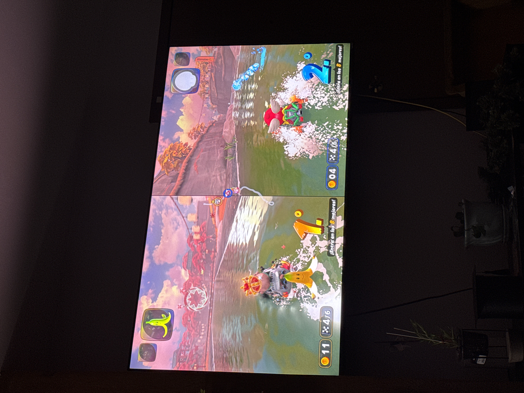The image shows a television screen displaying a split-screen view of a kart racing game. The top screen shows the player in second place, while the bottom screen shows the player in first place. The race is set on a water-themed track with pink and red decorations. The game interface shows the number of coins collected and the current lap out of six. The background includes a dark wall and some indoor plants. The image was taken indoors.