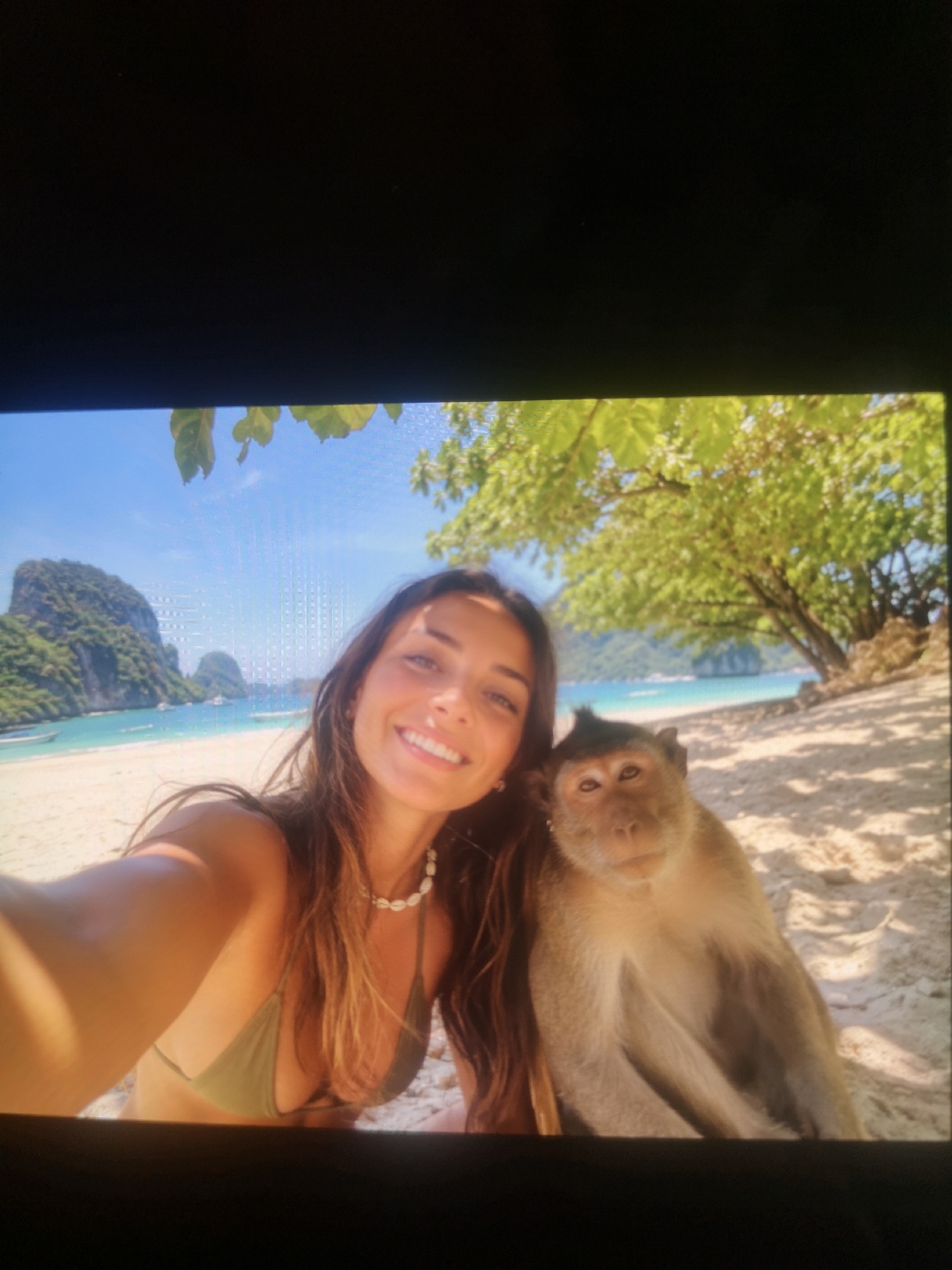 A woman is taking a selfie with a monkey on a beach. The woman has long brown hair and is wearing a green bikini top and a shell necklace. The monkey is light brown and is sitting next to her, looking at the camera. Behind them is a tropical beach with white sand, blue water, and lush green islands. The sky is blue and clear. A tree is overhead, partially obscuring the top right of the frame. Location: Unknown, Unknown.