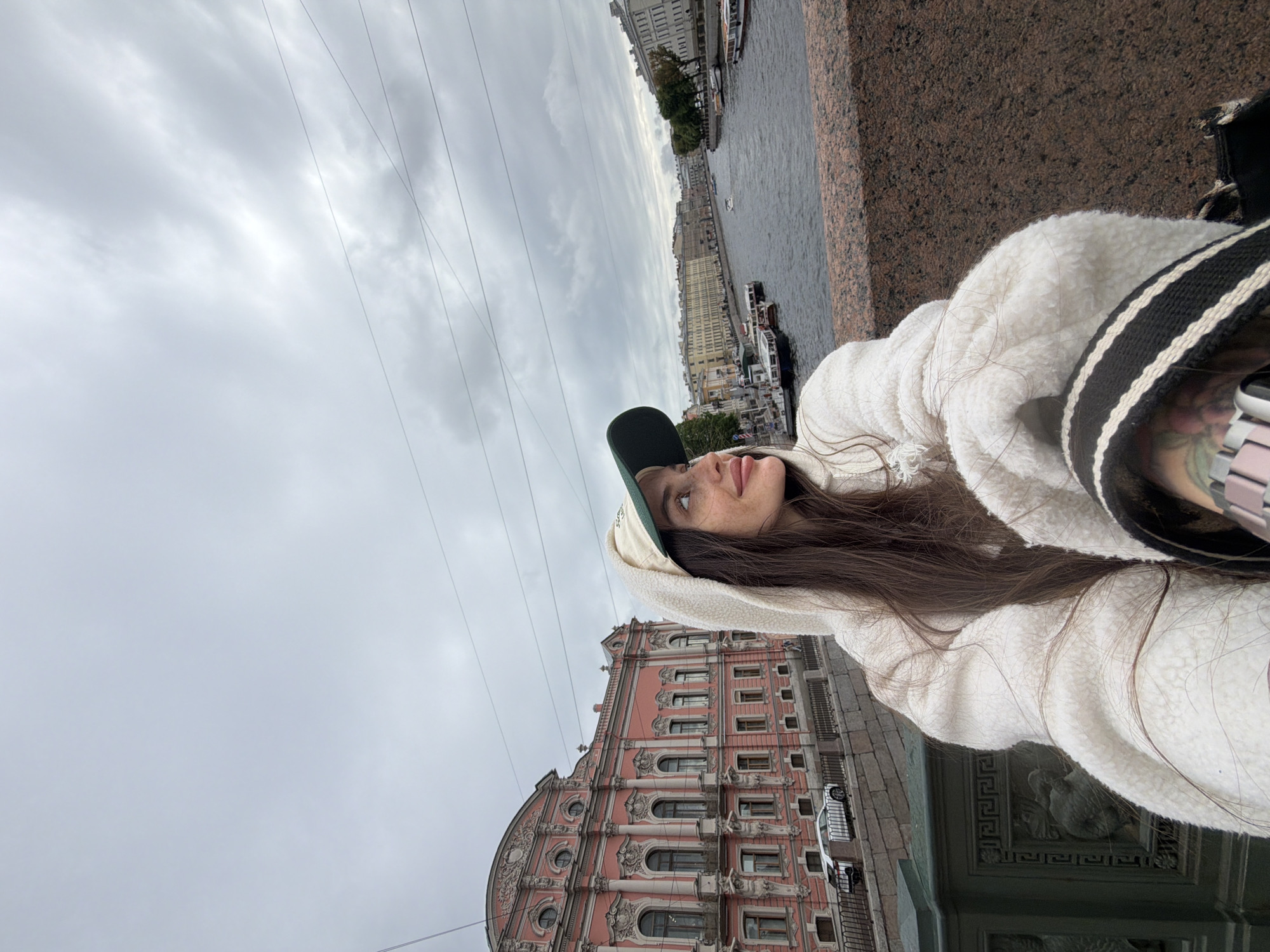 In Saint Petersburg, Russia, a young woman with long brown hair, wearing a white fuzzy hoodie and a green baseball cap, is taking a selfie from a bridge overlooking a canal. She is looking off to the side, with a smile on her face. The canal is lined with buildings, and there are several boats on the water. In the background, a large pink building with ornate architecture stands out. The sky is overcast and the overall atmosphere is cool and grey.
