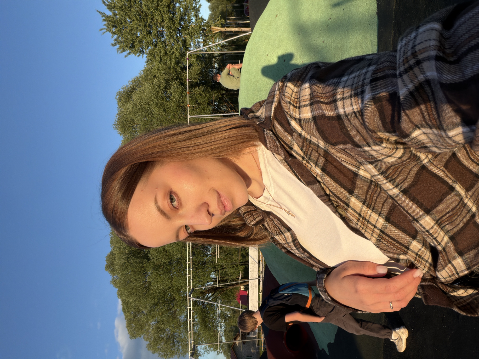 A young woman with light brown hair is centered in the foreground, wearing a white shirt and a brown plaid flannel. She is looking at the camera. In the background, there's a playground with green surfaces and metal structures. There are other people present, including at least one child. The sky is visible in the background and is clear blue. The setting appears to be outdoors in a park-like area. The geographic location, Sin'kovo, Russia, cannot be independently verified from the image.