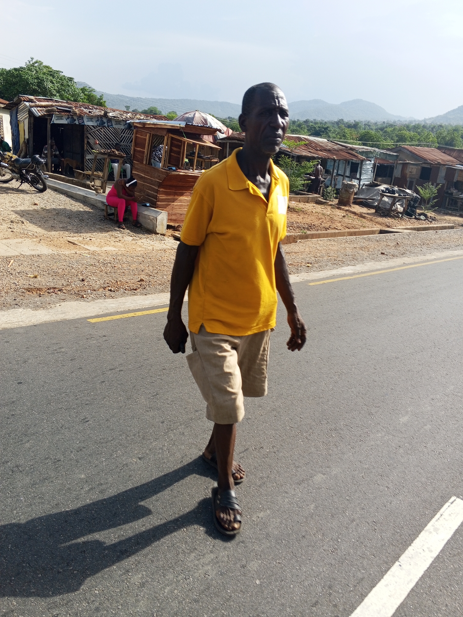 An older man, appearing to be of African descent, walks on a paved road under bright sunlight. He is dressed in a yellow polo shirt, light-colored shorts, and dark sandals, casting a prominent, elongated shadow on the asphalt, indicating the sun is not directly overhead. The road features a solid yellow line near the shoulder and a white line further out.
Adjacent to the road is a dusty, unpaved shoulder. In the mid-ground, a person in a bright pink outfit is seated on a concrete barrier, looking down. Behind the barrier, several rustic structures with corrugated metal roofs and wooden frames line the roadside, suggesting shops or stalls. Posters or advertisements are visible on the side of one structure, though the text is illegible. A black motorcycle is parked nearby, and a dog can be seen near one of the stalls.
The backdrop features lush, green hills under a clear, somewhat hazy sky. The scene captures a daytime moment of daily life in Adankolo, Nigeria, conveying a sense of warmth and activity in a rural or semi-urban setting.