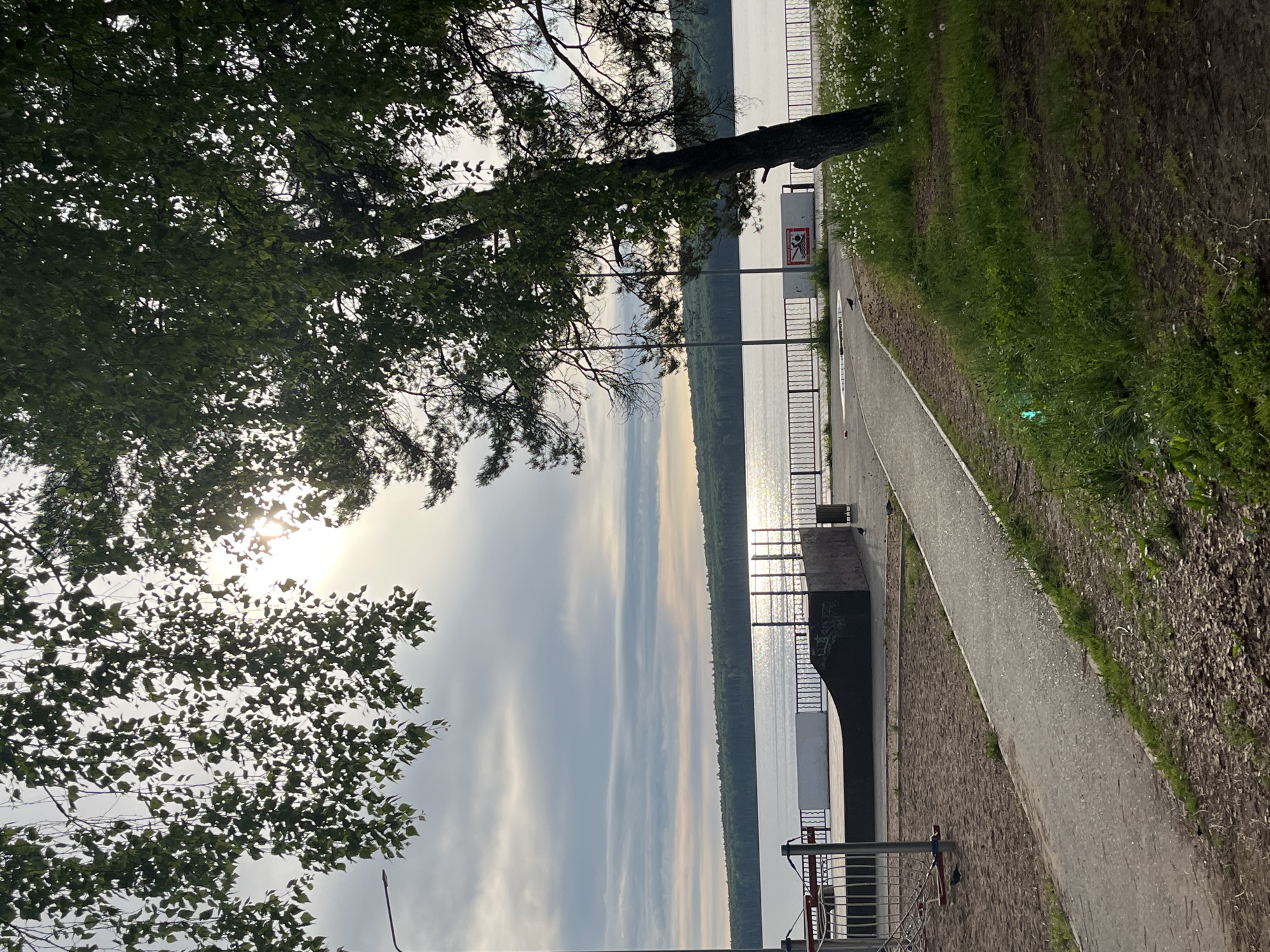 The image shows a path along a body of water, likely a river or lake, in Chaikovsky, Russia. The path is paved and bordered by grass and trees on one side and a metal railing overlooking the water on the other. The water surface reflects the sky, suggesting calm conditions. The sky is partly cloudy. Trees frame the image on the left side, partially obscuring the sun. There is signage next to the path and the water. A structure with a concrete base and metal railings is visible along the water's edge. There are also some grassy area and a few small white flowers next to the path.