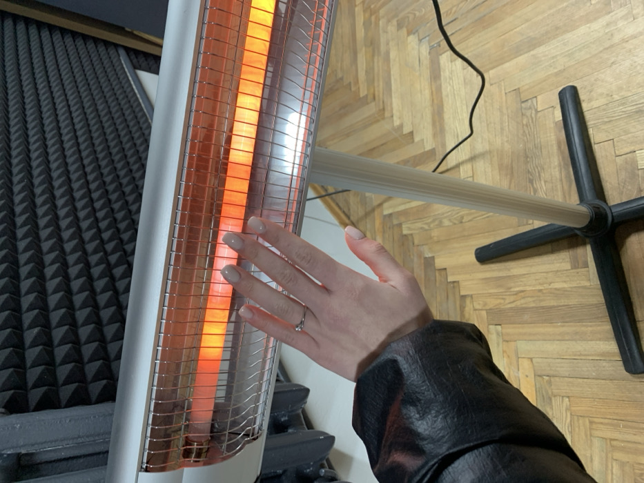 A forensic examination of the image reveals an indoor scene, located in the city of Kyiv, Ukraine. The primary subject is a radiant electric heater and a human left hand.
The heater is a silver-toned, horizontal unit with a protective metal grille, displaying two actively glowing, orange-red heating elements. It is supported by a light-colored vertical pole that extends downwards to a black, four-legged cross-base resting on the floor. A black electrical cord is visible trailing from the heater towards the right.
A light-skinned left hand is positioned in the foreground, palm facing the heater's glowing elements, suggesting an act of warming. The hand displays manicured fingernails with light-colored polish. A silver-toned ring, featuring a small, clear stone, is worn on the ring finger. The person's forearm is partially visible, covered by a black, textured sleeve, consistent with a leather or faux leather jacket.
In the background, a wall is partially covered with dark gray or black acoustic foam panels, characterized by a uniform pyramidal pattern. To the far left, a dark, ribbed object, possibly a cast-iron radiator or piece of furniture, is partially visible. The floor is constructed of light-brown wooden parquet, arranged in a herringbone pattern.