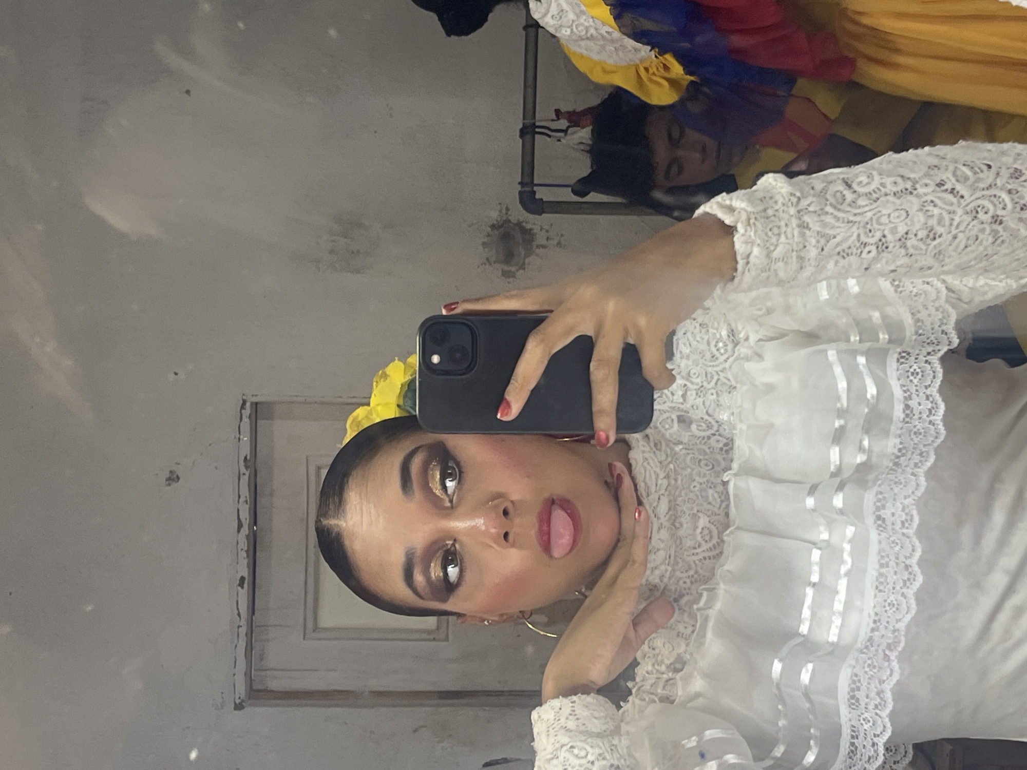 A young woman with dark hair pulled back and elaborate gold eye makeup is taking a selfie in a mirror. She is wearing a white lace and ruffled top. Her tongue is sticking out, and her left hand is resting on her chin. A yellow flower is tucked into her hair. In the background, a rack of colorful clothing is visible, and another person appears to be partially obscured. The setting appears to be a backstage area or dressing room, possibly within a theater or performance venue in Porlamar, Venezuela. The walls are textured and appear somewhat worn.