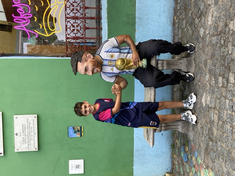 A young boy in a blue and pink soccer jersey shakes hands with a statue of Lionel Messi. Messi is wearing an Argentina jersey, black pants, and holding the World Cup trophy while sitting on a bench. They are in front of a green and blue painted wall, with historical plaques and a neon sign visible. The ground is cobblestone.