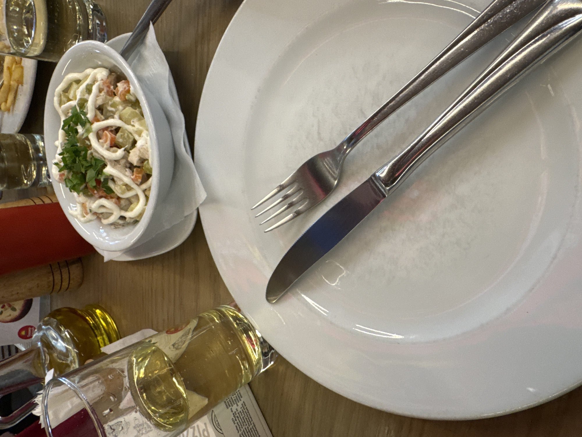 The image shows a tabletop scene, possibly in a restaurant in Yerevan, Armenia. A white plate with a fork and knife placed on it dominates the right side of the frame. To the left, there is a white bowl filled with a salad, likely Olivier salad, garnished with parsley. Other items on the table include a bottle of oil, a menu, and what appears to be a beer glass with some liquid. The lighting appears to be natural. The overall impression is that of a meal setting in a casual environment.