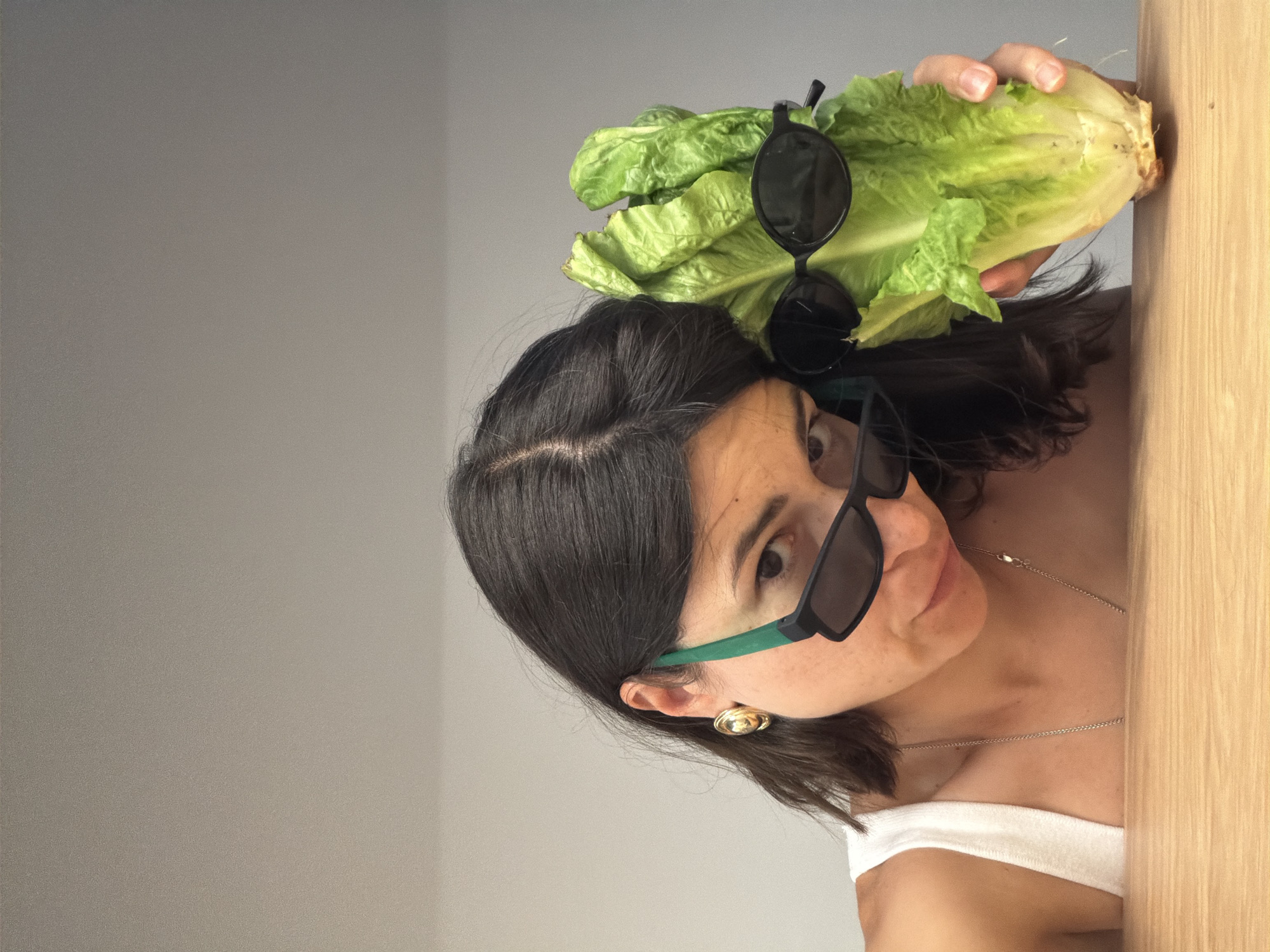 A woman is holding a head of romaine lettuce, on top of which rests a pair of black sunglasses. She is wearing sunglasses with green frames. She has dark hair and is wearing a white top and gold jewelry. She is standing next to a wooden door frame. The background is a gray wall. The location is unknown; the claim about the city of Budva, Montenegro, cannot be verified from the image.