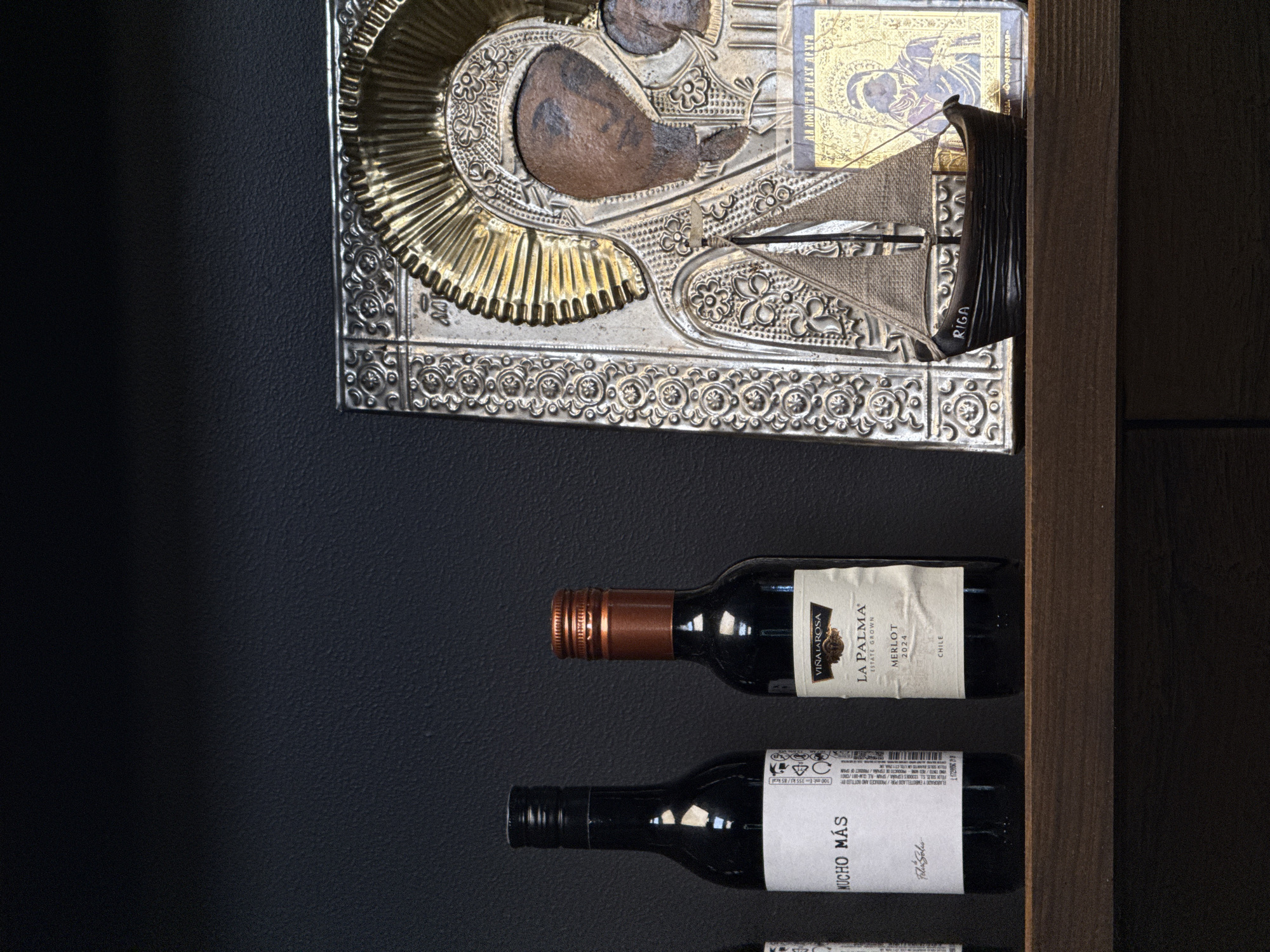 This is a close-up, eye-level shot of a wooden shelf in Tūja, Latvia. On the shelf are three wine bottles, a small model sailboat, and two religious icons. The leftmost wine bottle is black with a white label that reads "MUCHO MÁS." The middle bottle is also black with a white label that says "VIÑA LAROSA LA PALMA MERLOT 2024 CHILE." The third bottle is obscured by the sailboat. The model sailboat is dark brown with light brown sails and the word "RIGA" printed on its side. The two religious icons are ornate and appear to be made of metal and gold. The background is a dark, textured wall. The lighting is dim, with the objects on the shelf casting faint shadows.