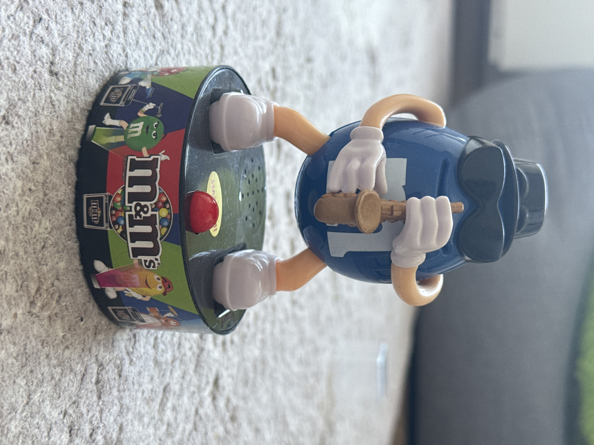 A blue M&M's character, wearing a black fedora and sunglasses, stands on a circular base, holding a small saxophone. The character has white gloves and white shoes. The base is decorated with various M&M's characters and the M&M's logo. A red button is visible on the top of the base. The object is placed on a light-colored carpet. The background is blurred, showing a hint of furniture. The scene is indoors, with soft lighting.