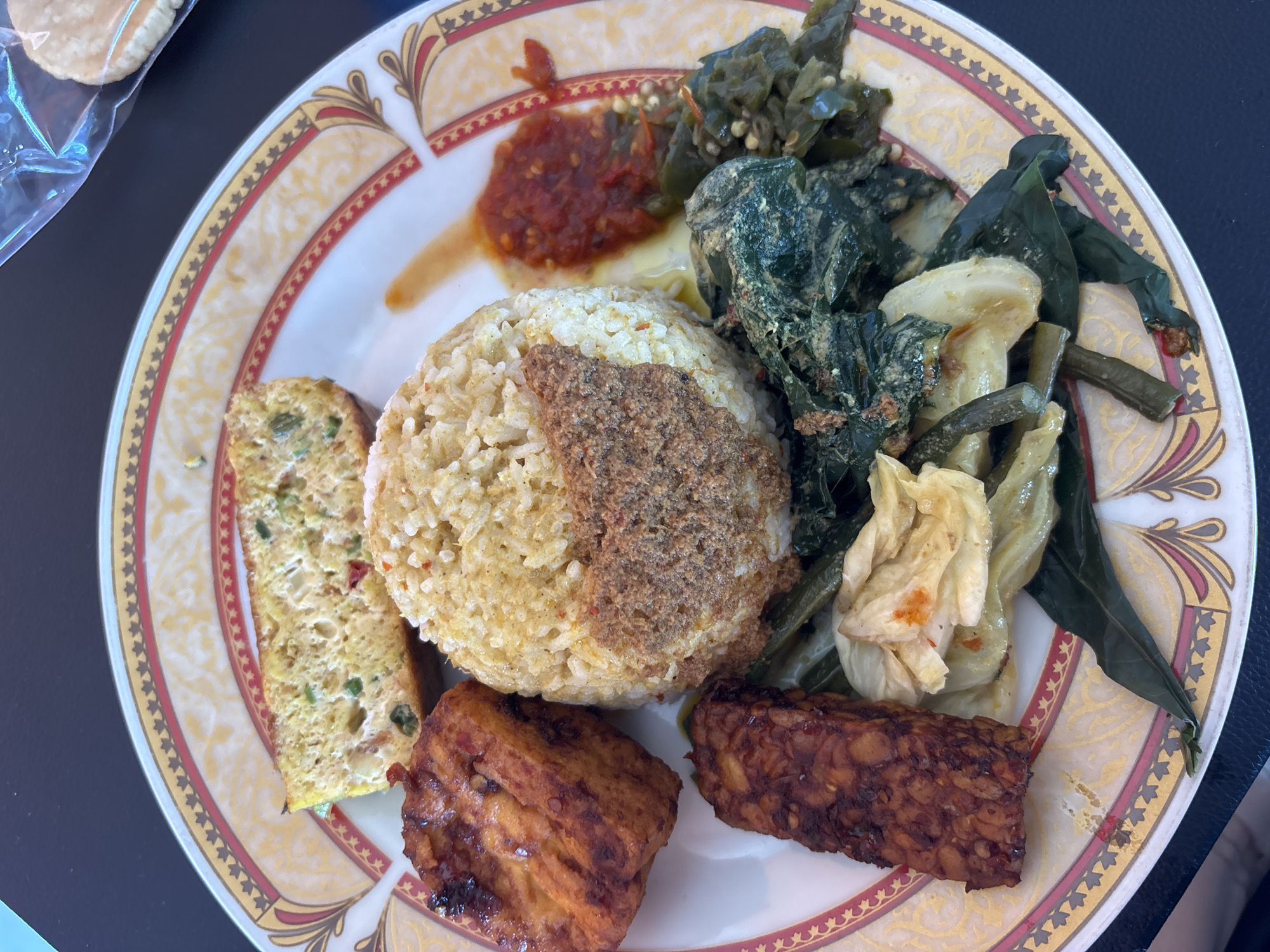 The image shows a close-up overhead view of a plate of Indonesian food, likely at a restaurant in Kuta Selatan, Indonesia, given the context of Indonesian cuisine. The plate, with a decorative rim featuring gold, red, and black patterns, contains a mound of rice topped with a dry, crumbly seasoning. To the left of the rice are two pieces of fried tempeh, one larger and one smaller. Adjacent to the rice and tempeh are various cooked vegetables, including leafy greens and what appear to be long beans. A slice of what looks like savory omelet or frittata with visible green chilies and red specks is positioned at the top of the plate. A dollop of red chili paste (sambal) is also visible. The background is a dark, solid surface, possibly a table. The overall presentation suggests a prepared meal ready to be eaten.