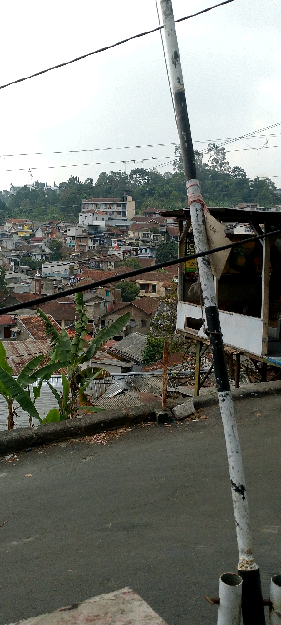 The image captures a hillside urban landscape in an Indonesian city, characterized by densely packed houses with red and gray tile roofs. A prominent, weathered black and white striped pole stands in the foreground, partially obscuring a small, open-air structure with a white facade and wooden supports. Overhead, a network of power lines stretches across the frame against a hazy, overcast sky. Lush green banana trees grow on the left side of the image, their large leaves reaching upwards. The foreground shows a sloping asphalt road.