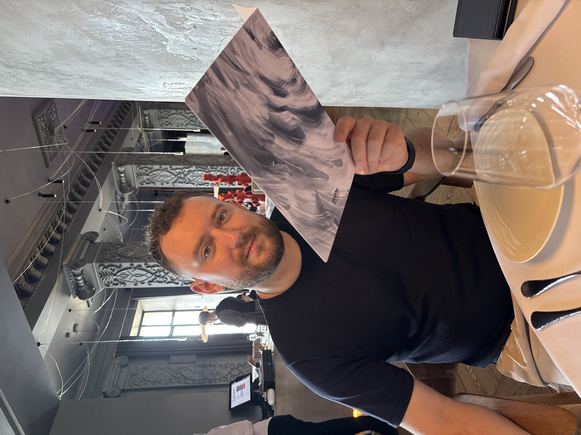 An indoor, slightly rotated image captures a man holding a menu in a restaurant or cafe in Krasnoyarsk, Russia. The main subject is a man with light skin, dark hair, and a short beard, wearing a black long-sleeved t-shirt and a black wristband on his left wrist. He looks directly at the camera with a neutral to slightly pleasant expression. He holds an open menu with a grey and white abstract, swirling pattern, featuring the text "BWFM" vertically printed in the bottom right corner.
In the foreground, a table covered by a white tablecloth displays a light-colored plate, a fork and knife, and a clear wine glass. The man is seated on a wooden chair.
The setting reveals an interior with ornate decorative molding on the walls and ceiling, contrasting with modern pendant lights suspended by thin wires. In the blurry background, other patrons or staff members are vaguely visible, along with a screen on a counter that partially shows the text "LifeSaver". Light emanating from a window suggests it is daytime. The scene depicts a casual dining or social event in a sophisticated setting.