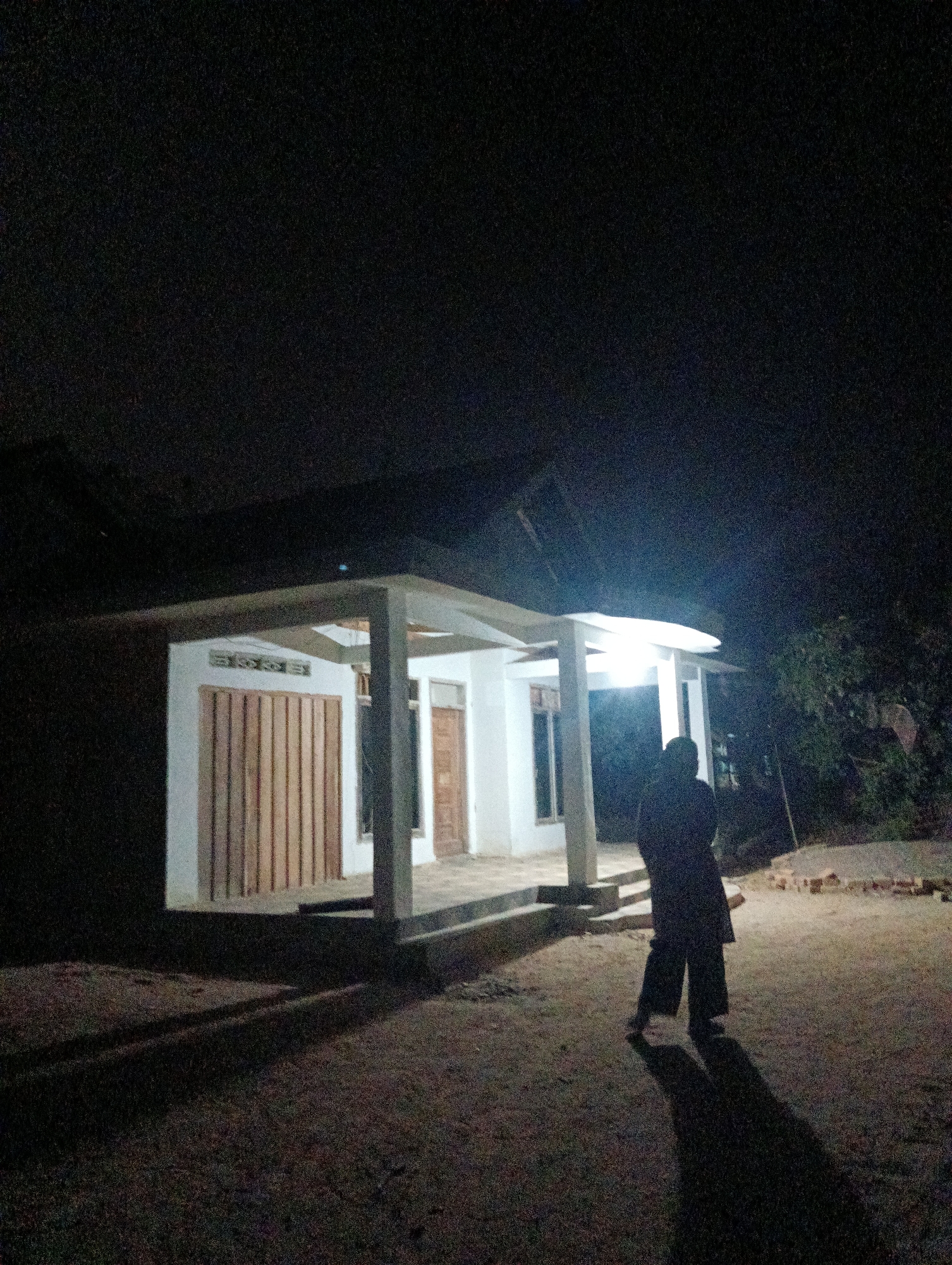 A person stands in front of a white house at night in Pematang Jaya, Indonesia. The house has a covered porch with supporting pillars and a light illuminates the front of the building. The person is silhouetted against the light and appears to be wearing dark clothing. The ground in front of the house is unpaved and appears to be dirt or gravel. Shadows are cast on the ground from the house and the person. Trees are visible in the background to the right of the house. The overall scene suggests a residential area in a rural or semi-rural setting.