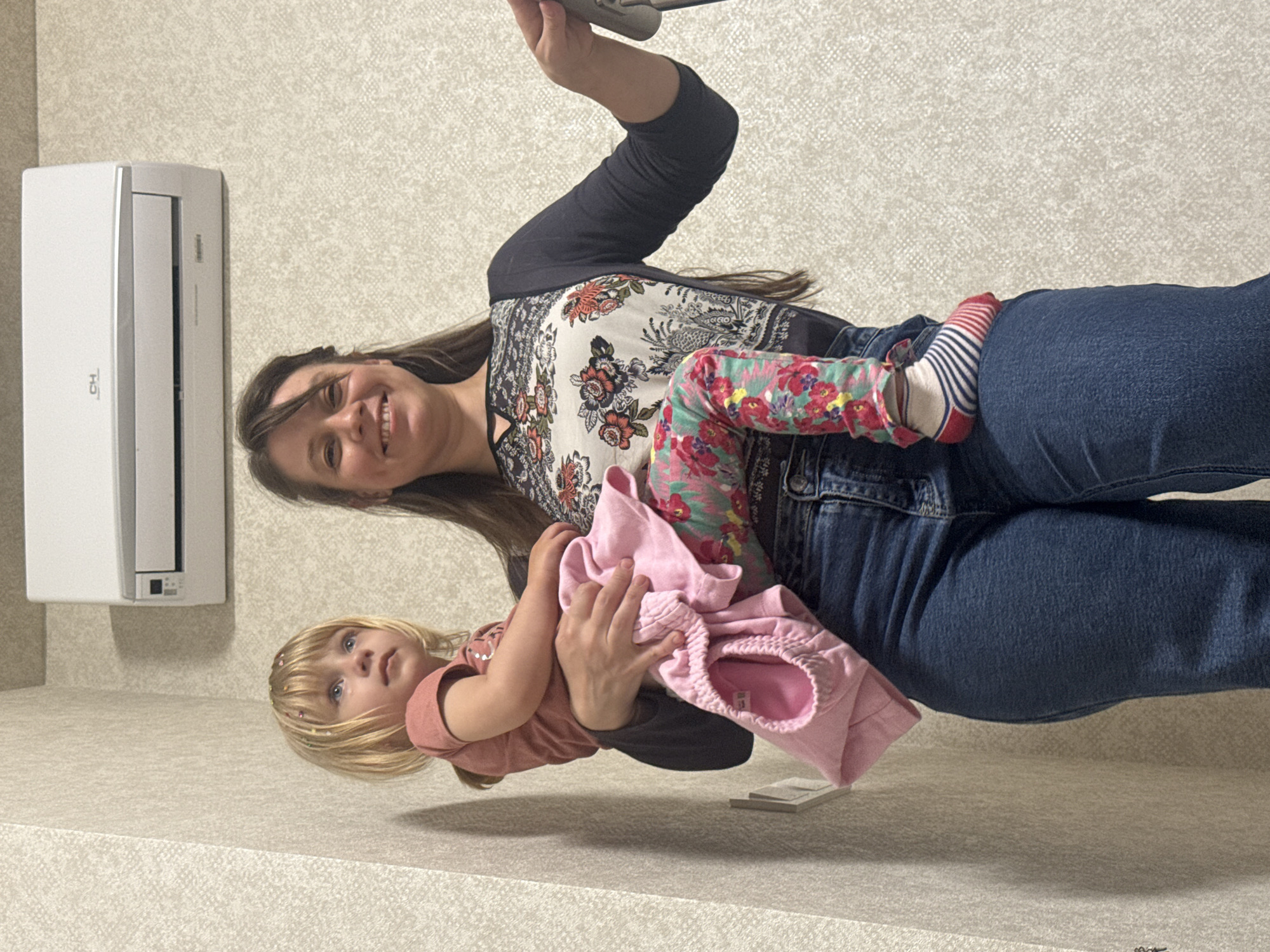 The image depicts a woman holding a young child. The woman is wearing jeans, a floral top, and a black cardigan. The child is wearing a pink shirt and floral pants with red and white striped socks. The woman is smiling. In the background is a wall-mounted air conditioner. The location is Unknown, Unknown.