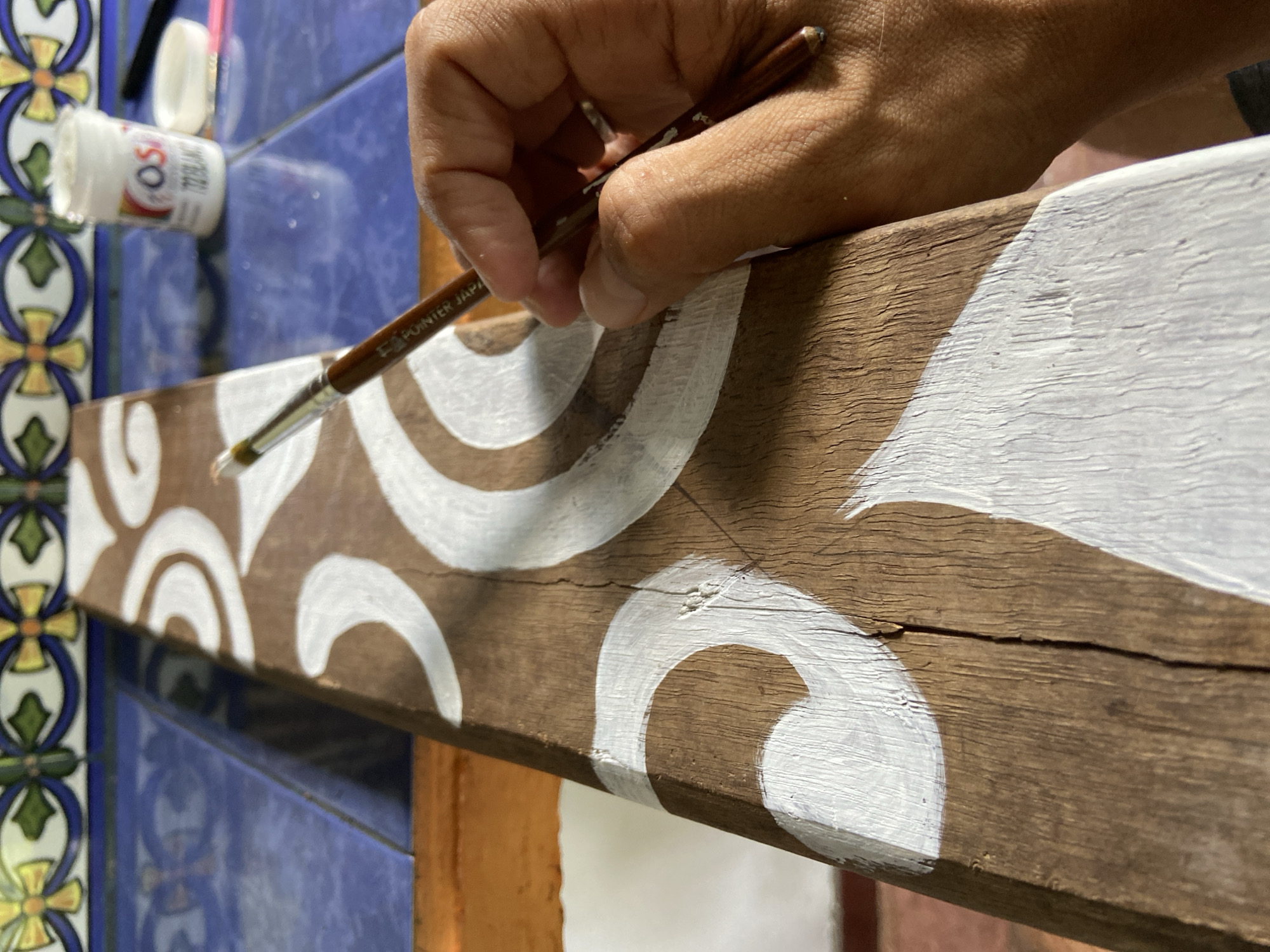 A close-up, eye-level shot captures a person's right hand actively painting white patterns onto a rustic wooden plank. The hand firmly grips a brown paintbrush, which bears the partial text "POINTER JAPA" (likely Japan) on its handle. The tip of the brush, laden with white paint, is poised above or making contact with the wood. The wooden plank, characterized by its natural grain and a prominent crack running its length, is adorned with swirling and curved white designs, some freshly applied with visible brush strokes and others appearing dry.
In the blurred background to the left, a white bottle of "OSCAR ACRILICAS 722 BLANCO" (white acrylic paint) rests on a vibrant blue tiled surface. These blue tiles, showing reflections, are bordered by decorative tiles featuring intricate yellow, green, and blue floral or geometric patterns. A subtle shadow falls across the painted plank. The scene suggests an artistic activity taking place in a well-lit environment, possibly indoors or in a covered outdoor area in Los Velásquez, Venezuela. The focus is on the intricate work of painting and the textures of the wood and paint.