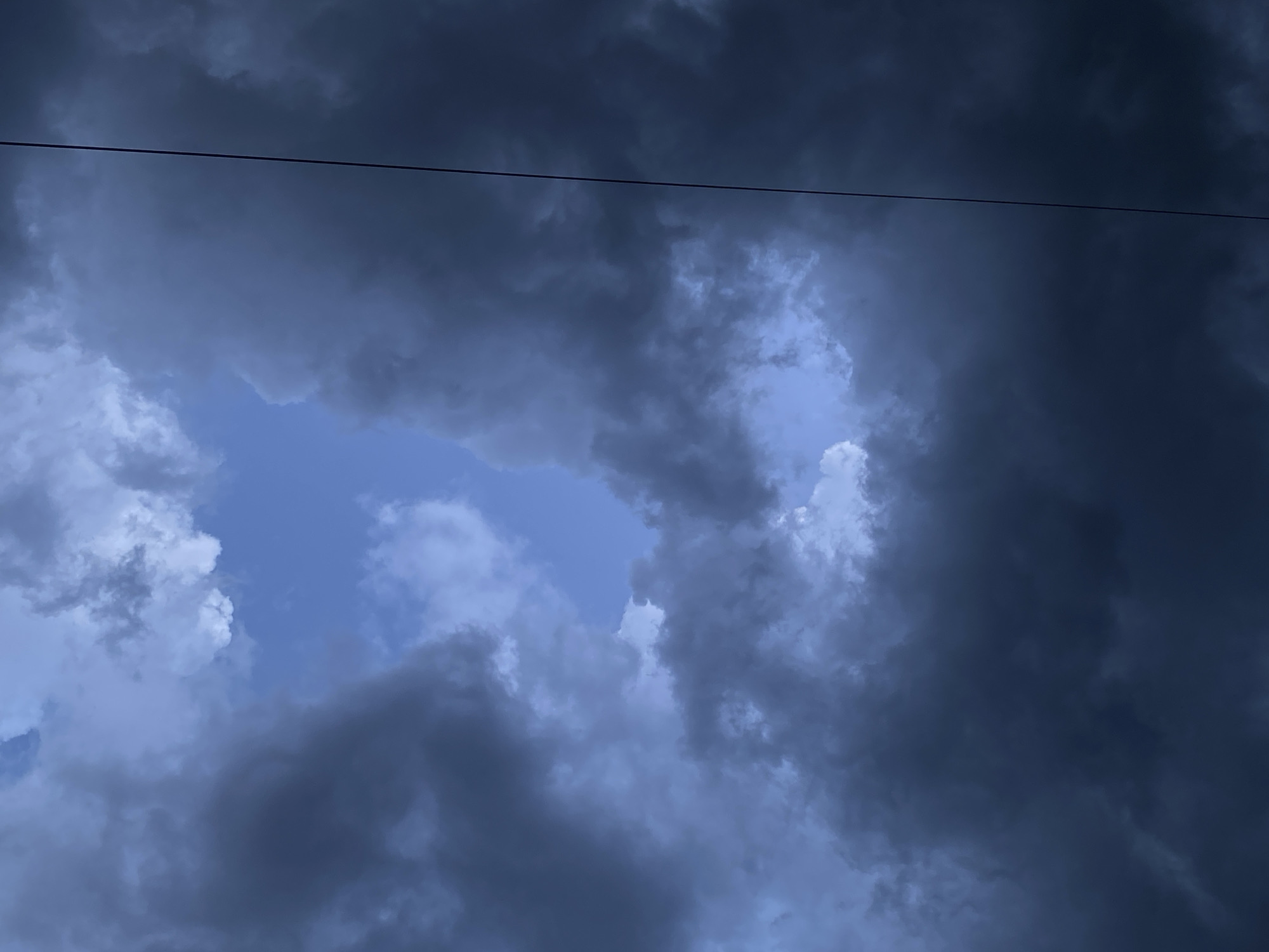 A low-angle, overcast shot of dark storm clouds with a small area of lighter blue sky visible in the center. A thin, dark wire stretches across the top of the frame. The photo appears to be taken outdoors. There are no people or objects present in the frame other than the sky and the wire.